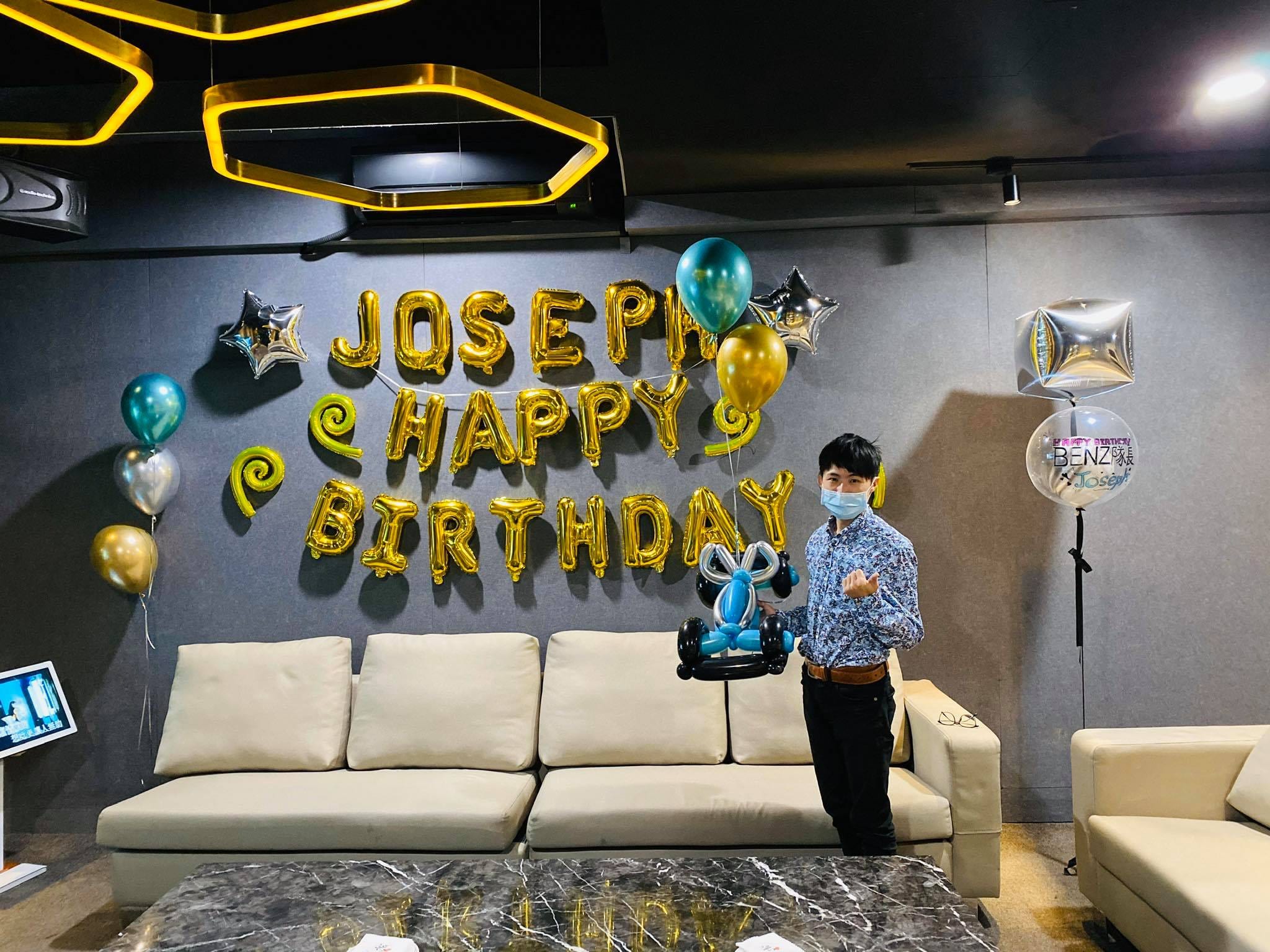 我拿著藍銀賽車造型氣球，站在金色 HAPPY BIRTHDAY 字母牆前