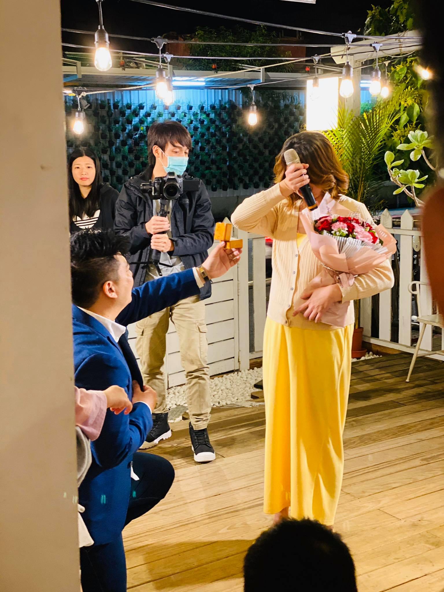 穿深藍西裝的男士單膝跪地遞上戒指，女士捧花拭淚