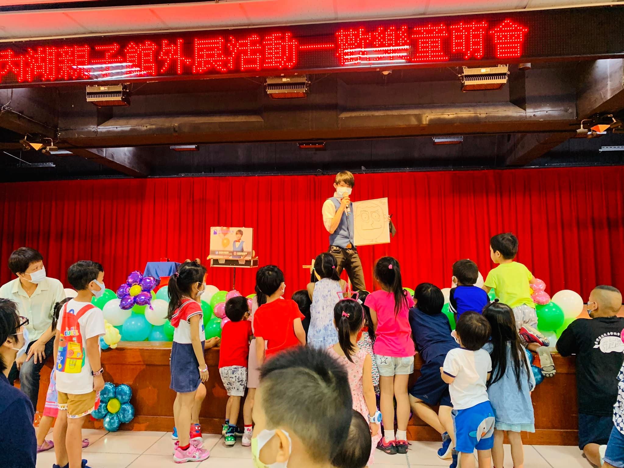 戴口罩的魔術師在紅色布幕舞台上拿畫板表演，孩子圍在台前