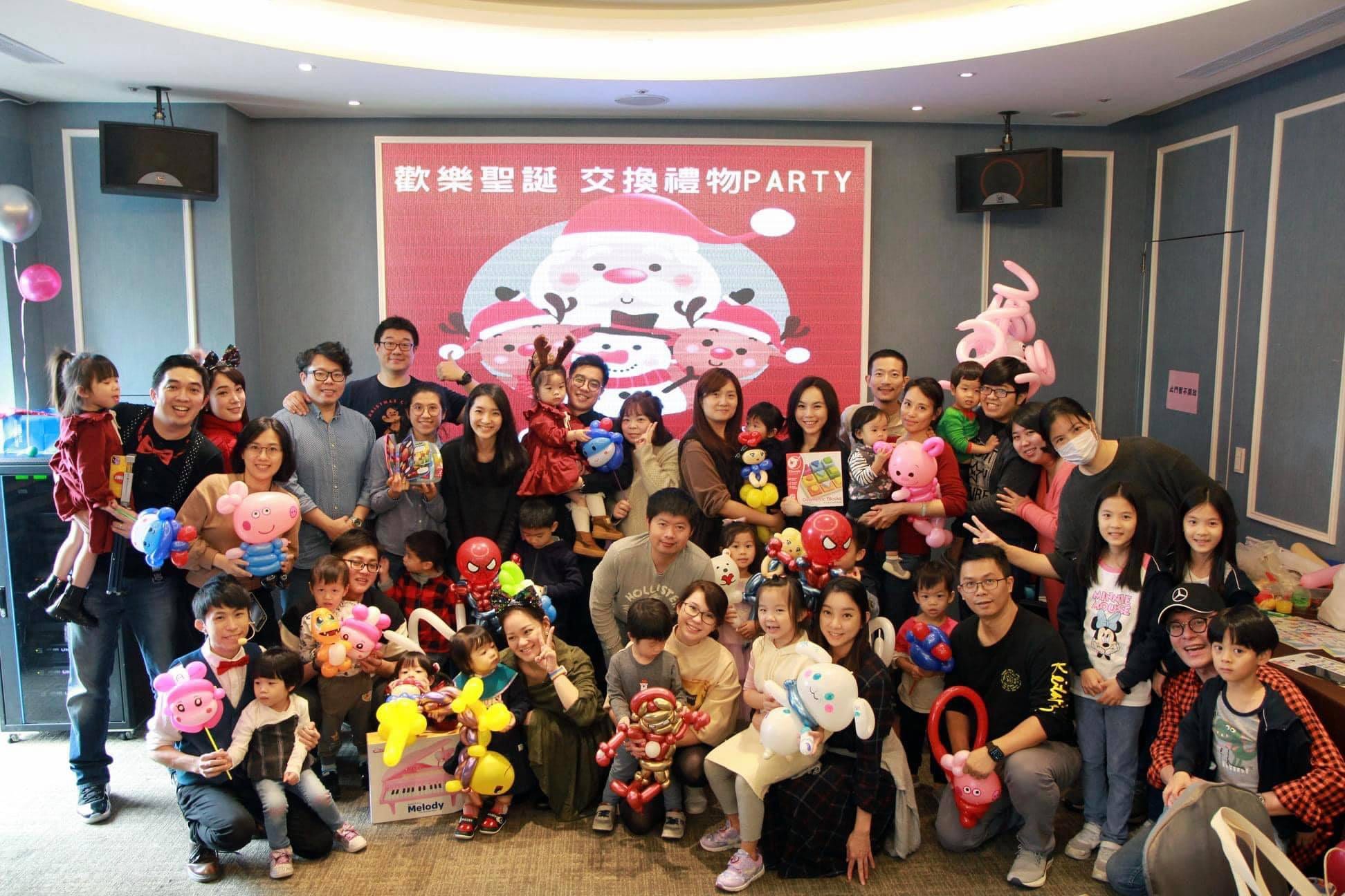 小屁孩聖誕派對大合照，大人小孩拿著造型氣球站在交換禮物背板前