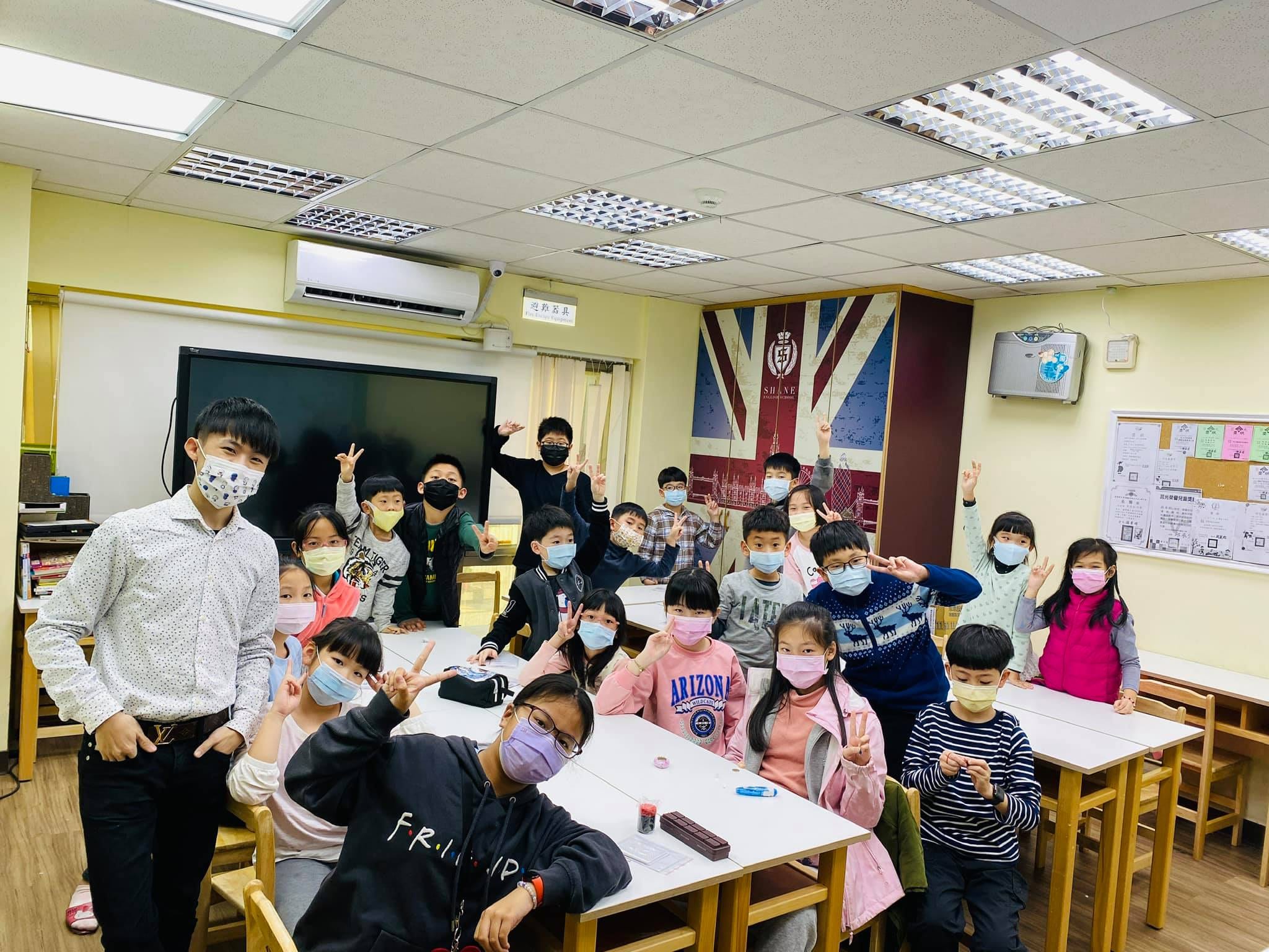 掛著英國國旗的教室裡，小朋友和柯男比手勢合照