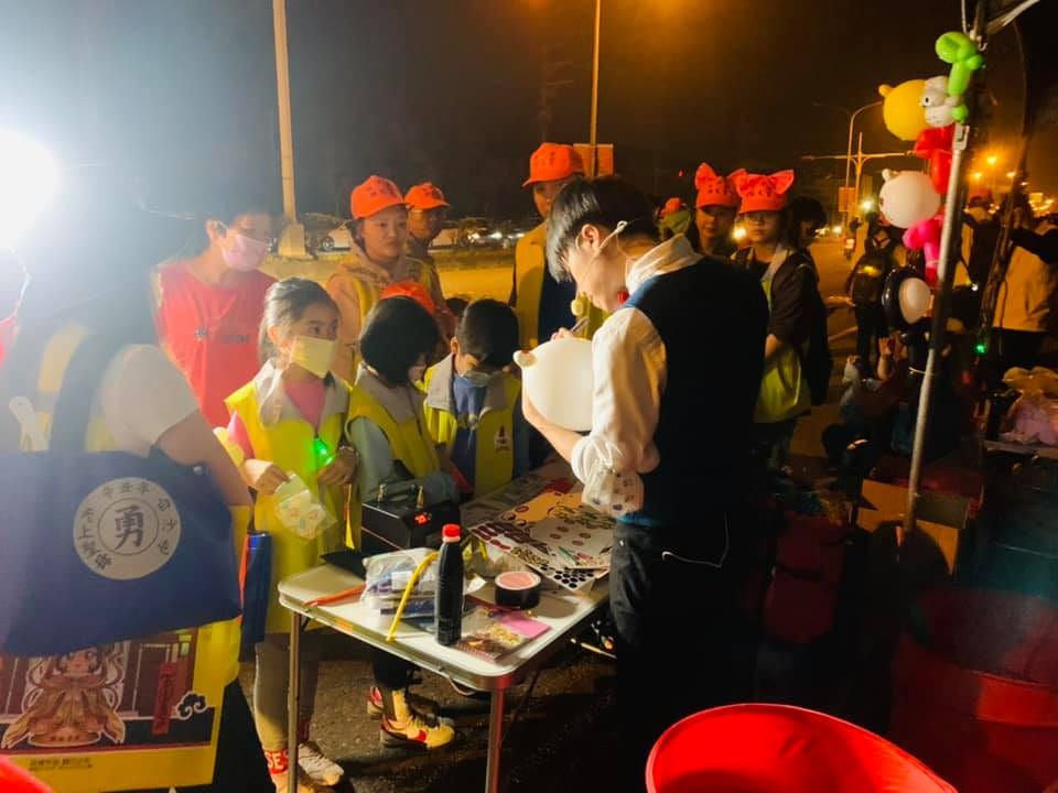 夜晚路燈下，柯男在小桌前折氣球，進香民眾圍在攤位邊