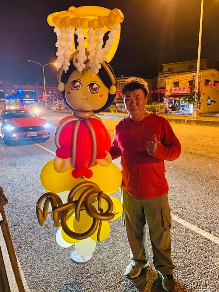 夜晚路邊，穿紅衣的大哥站在氣球媽祖旁比讚