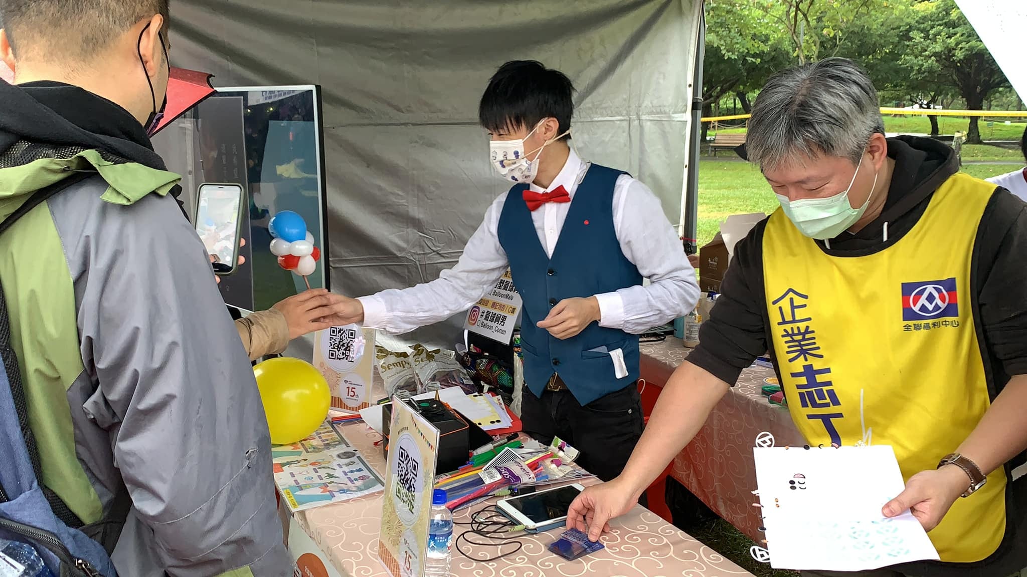 柯男在攤位遞出造型氣球，穿黃色企業志工背心的工作人員協助