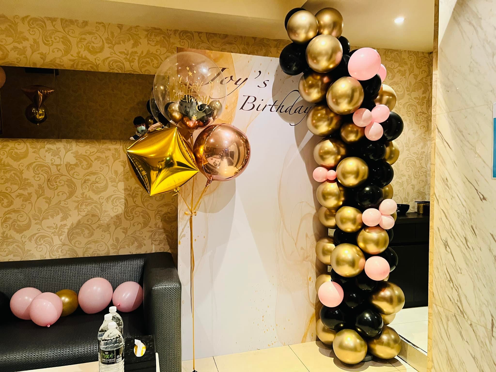 寫著 Joy's Birthday 的白色客製背板，右側黑金粉不規則氣球柱，左側金色星星、玫瑰金圓球與波波球空飄氣球束