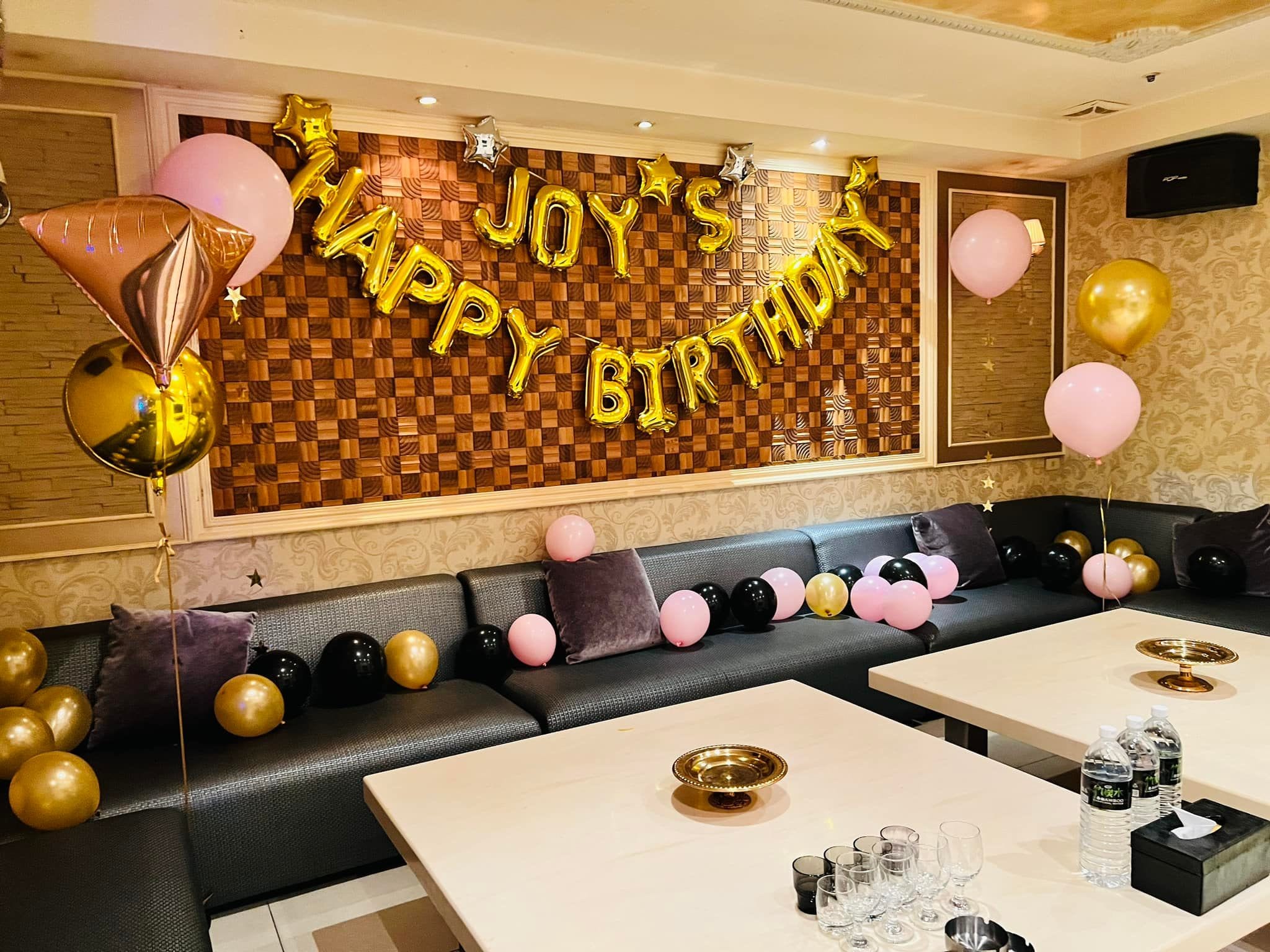包廂木紋格子牆上的金色 JOY'S HAPPY BIRTHDAY 字母氣球，點綴銀色和金色星星，沙發上鋪滿粉黑金氣球，左側玫瑰金鑽石與金色圓球空飄氣球
