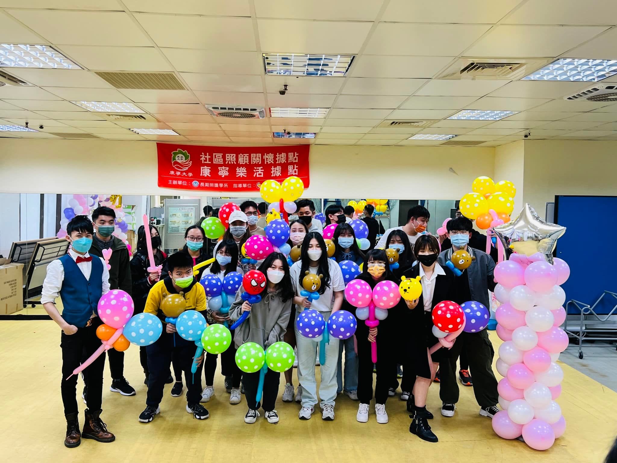 教室裡掛著社區照顧關懷據點康寧樂活據點紅布條，二十多位大學生拿著點點氣球棒與動物氣球合照，右側是粉白氣球柱