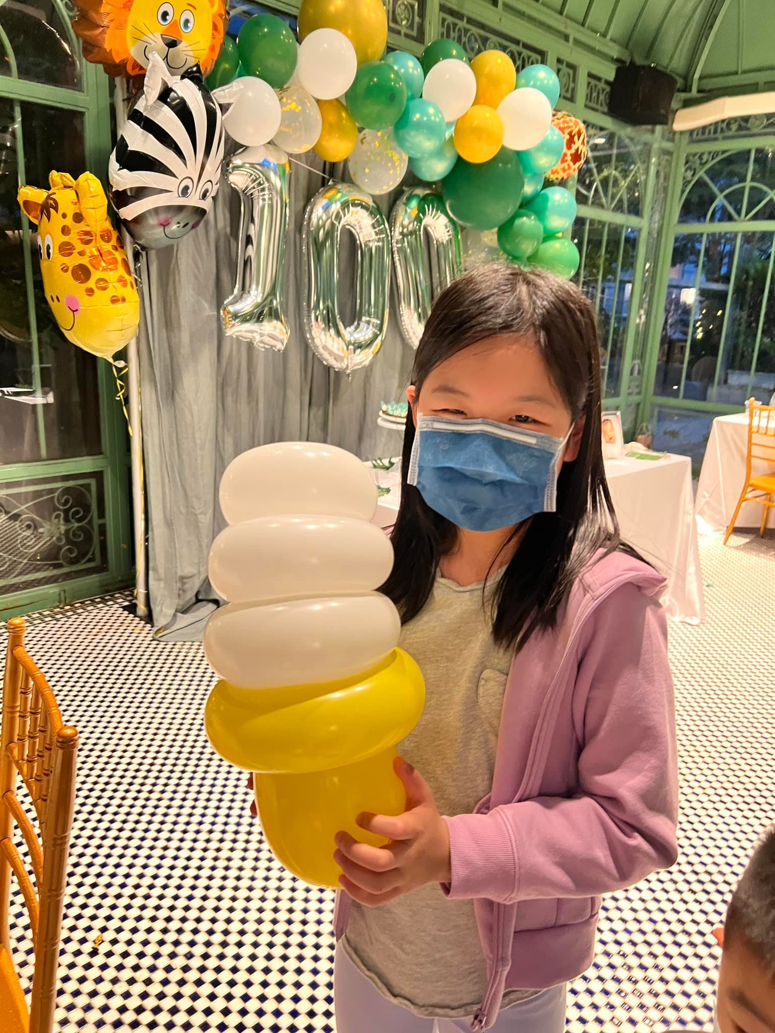 小女孩拿著白色和黃色的冰淇淋甜筒氣球，背後是銀色數字 100 氣球與獅子、斑馬、長頸鹿鋁箔氣球
