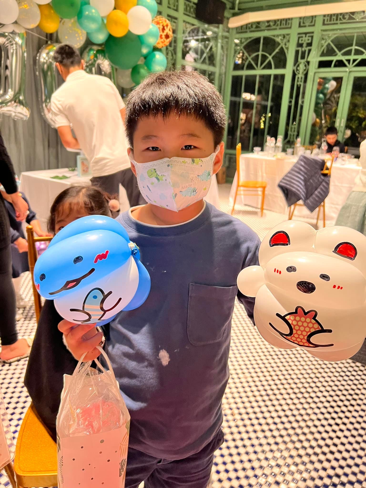 小男孩一手拿藍色小鯊魚氣球、一手拿白色小熊氣球，後方是玻璃溫室風格的餐廳