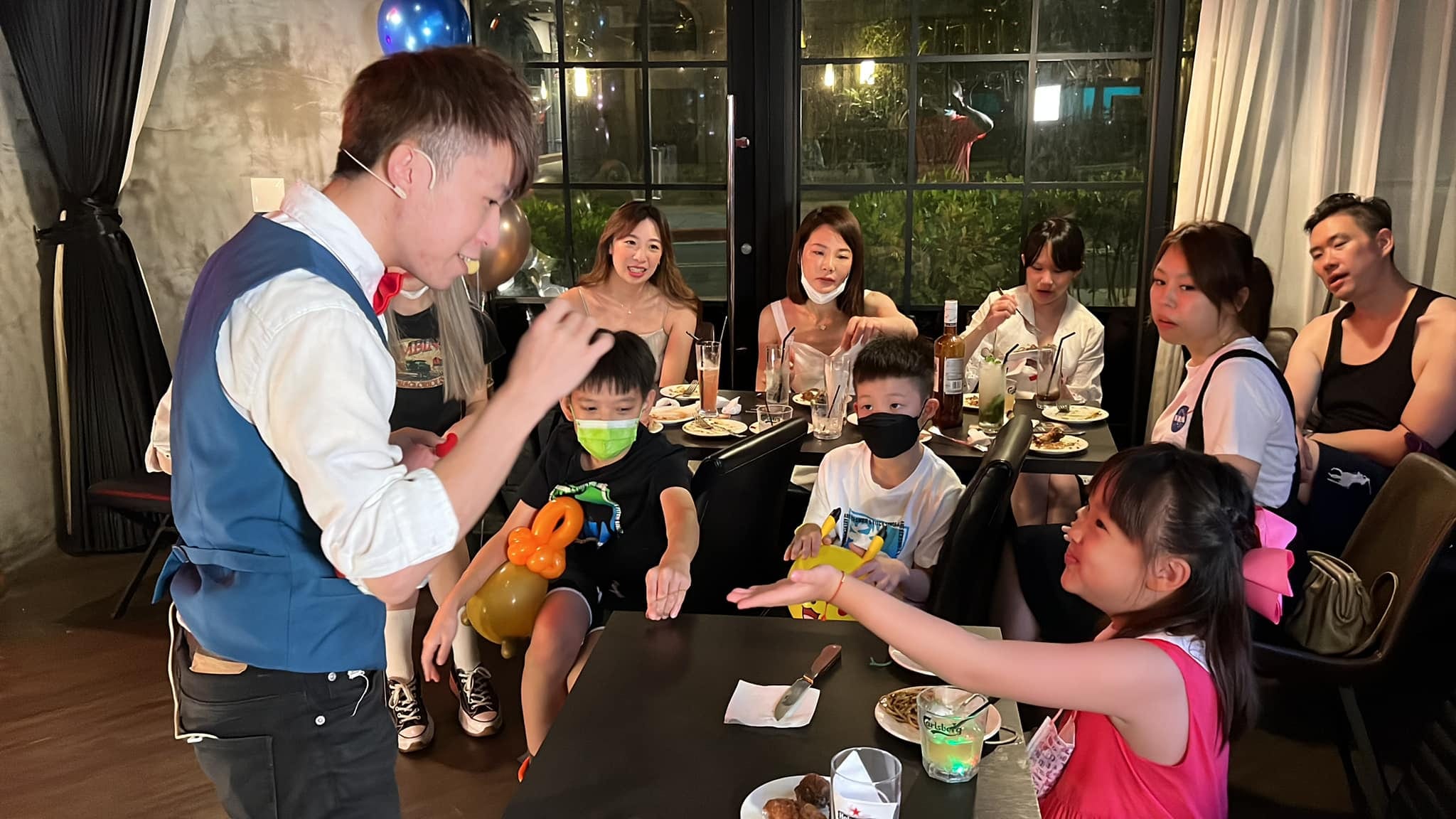柯男站在餐桌旁變魔術，小女孩伸手、大人們看得很專心