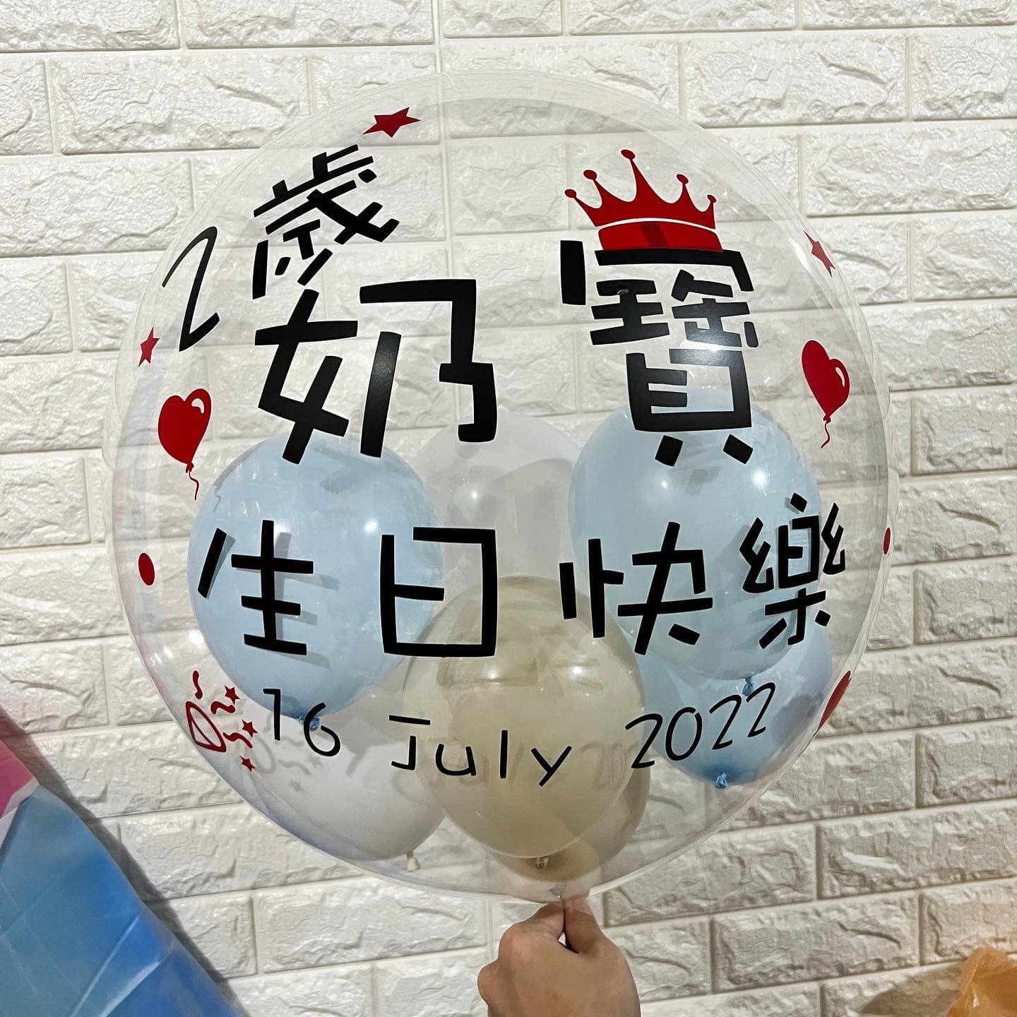 透明波波球上寫著「2歲奶寶生日快樂 16 July 2022」，裡面裝著淺藍、白色與奶茶色小氣球
