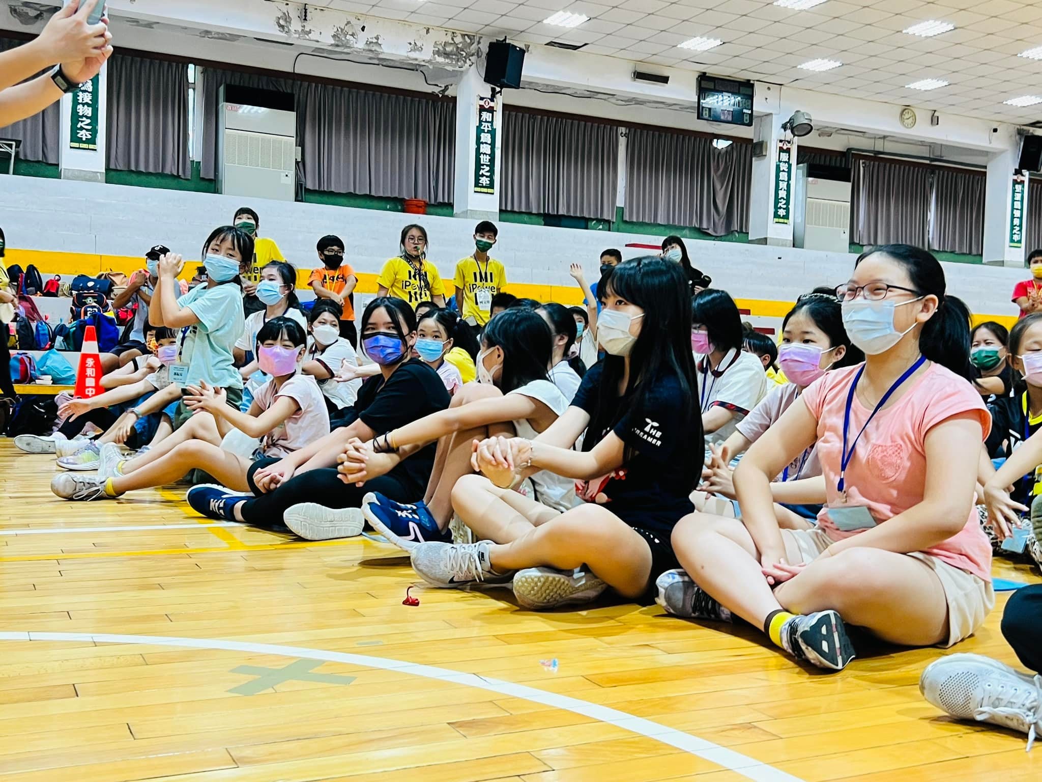 體育館木地板上，戴名牌的孩子們盤腿坐著看表演