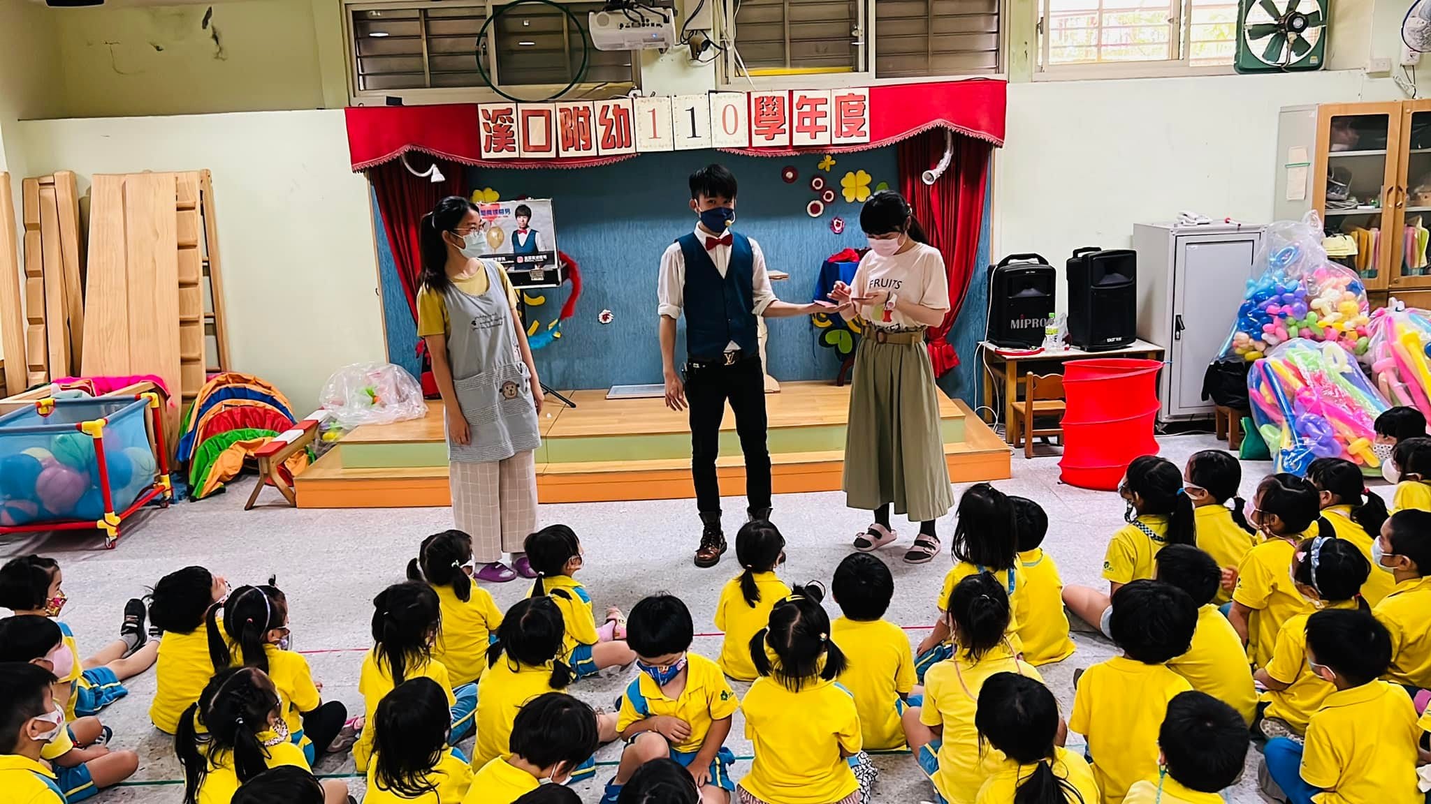柯男和兩位老師站在幼兒園舞台前，台下孩子整齊坐好