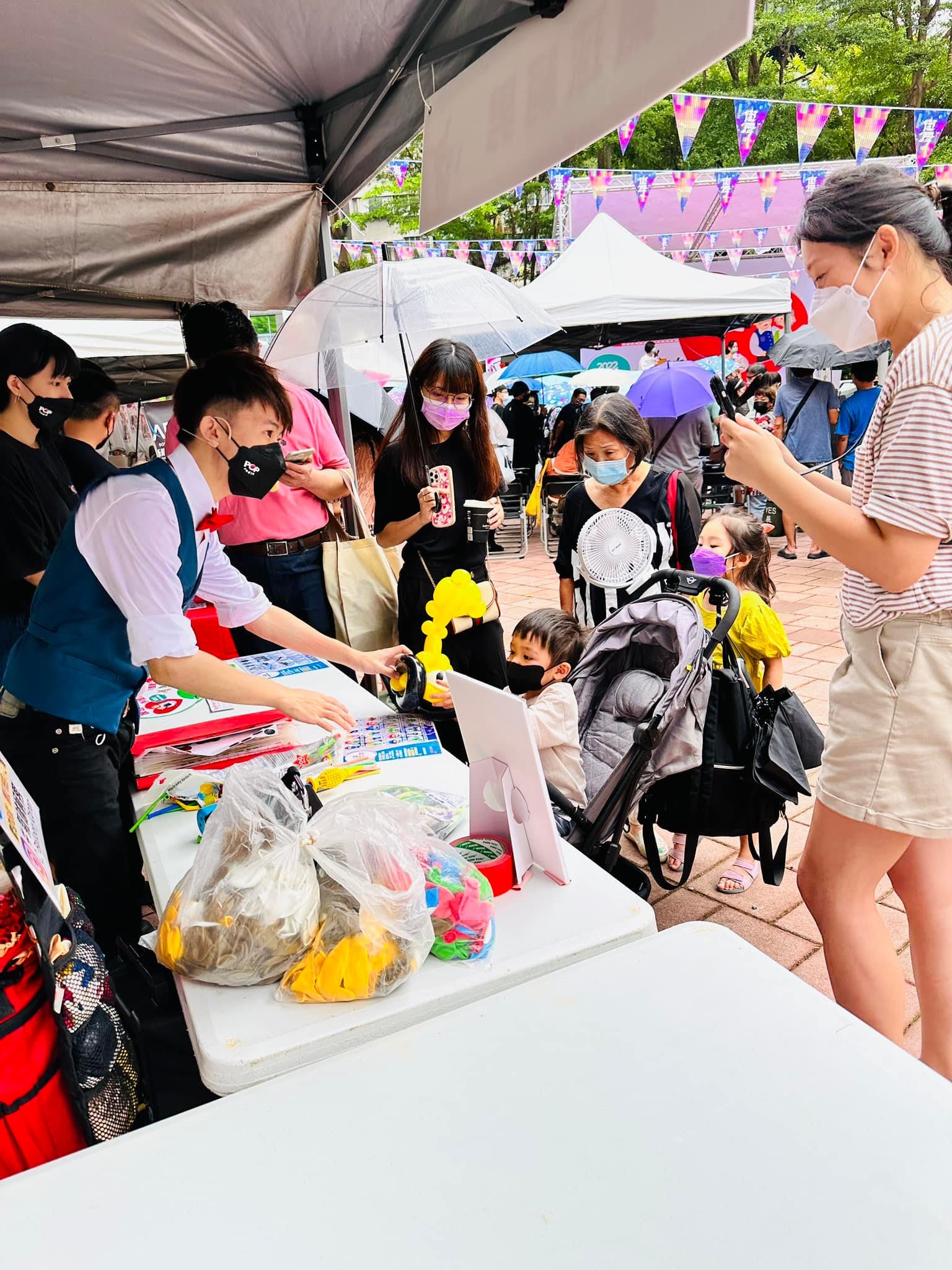 家長推著嬰兒車在氣球攤位前等待,柯男正在桌邊遞出黃色氣球