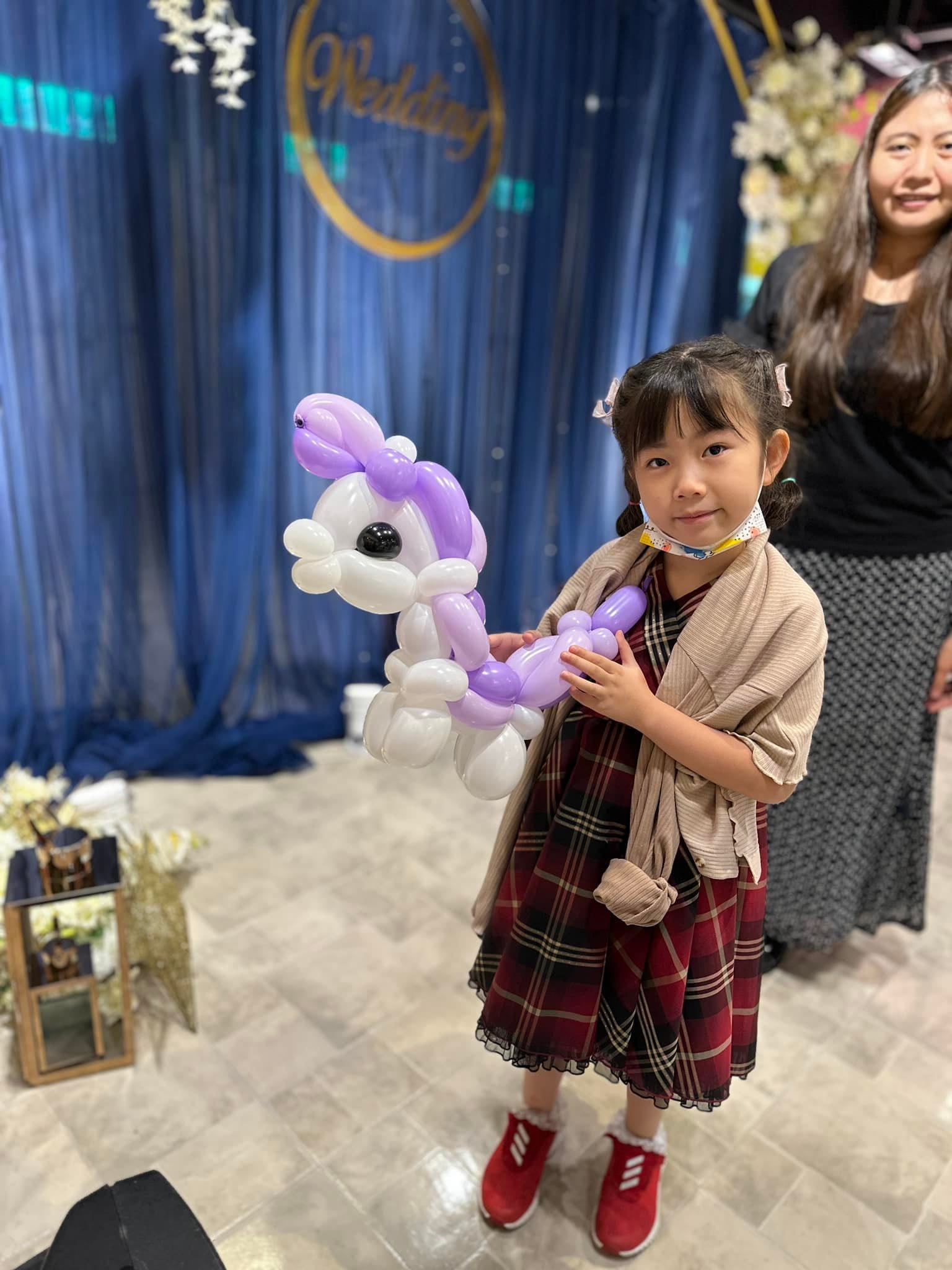 穿格子洋裝的小女孩抱著紫色鬃毛的白色小馬氣球,背後是 Wedding 背板