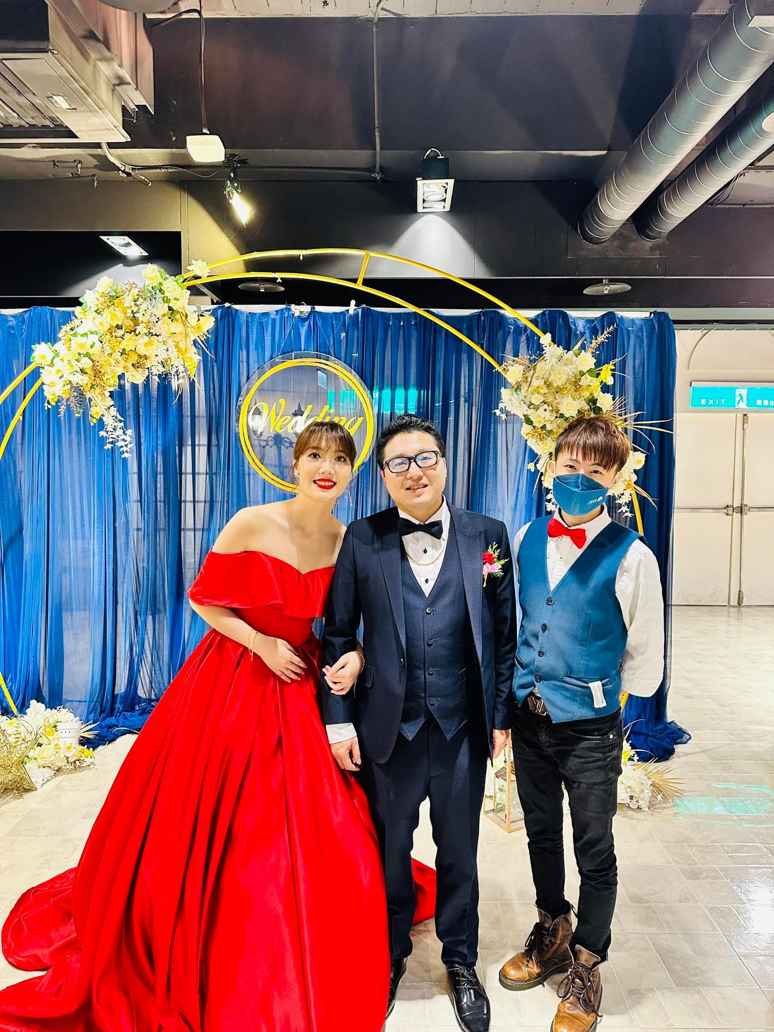新娘穿紅色禮服、新郎穿深藍西裝,與柯男在藍色布幔與金色圓拱的婚禮背板前合照