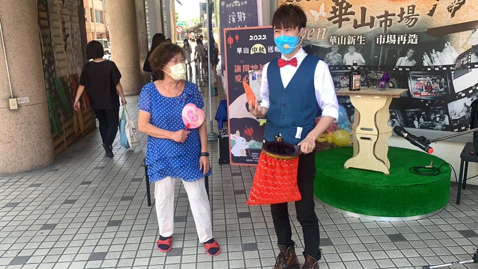 柯男拿著紅色魔術袋,旁邊穿藍色碎花上衣的長輩拿著粉紅色道具