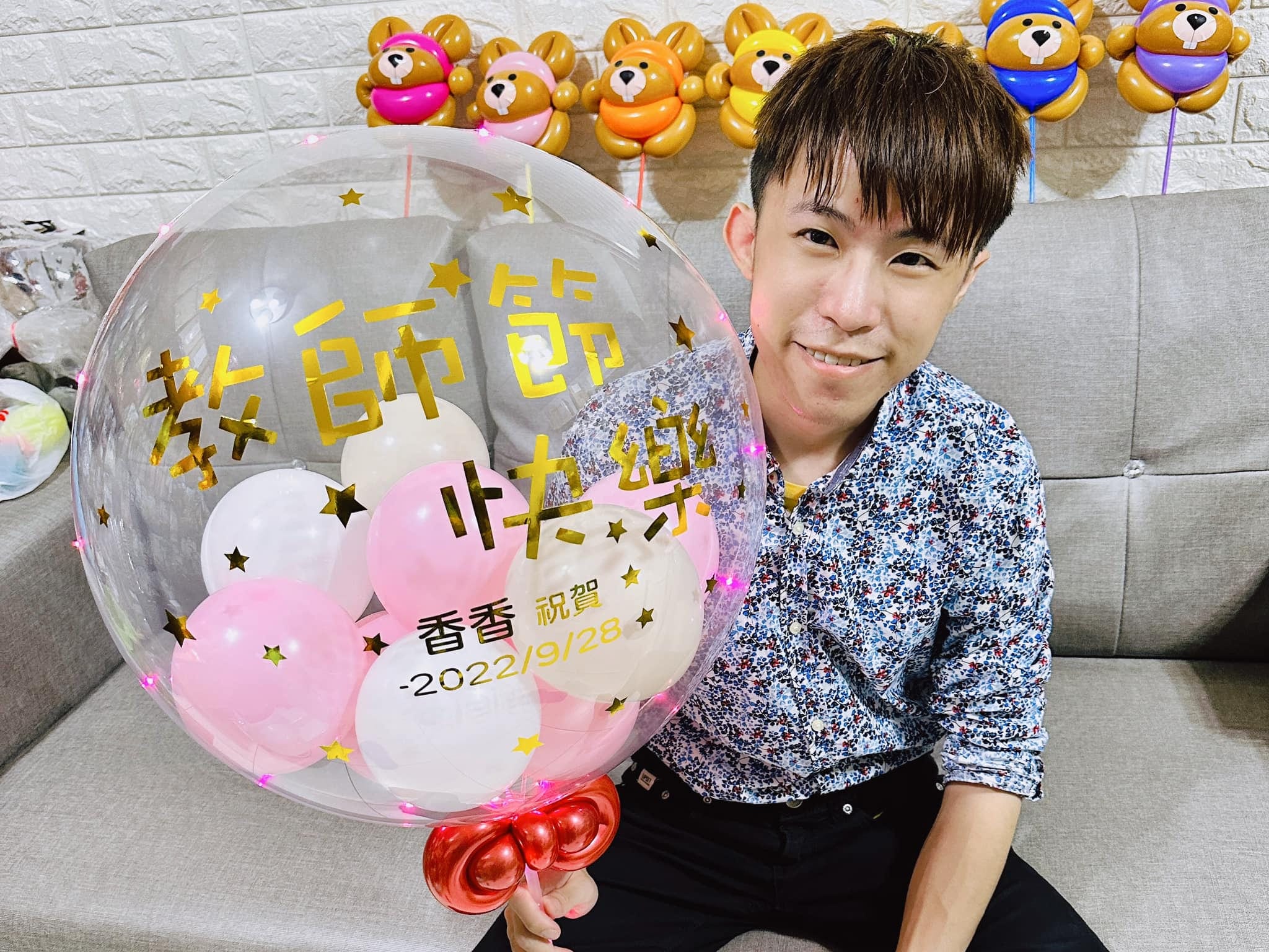 我坐在沙發上拿著透明波波球，球面金字寫著教師節快樂、香香祝賀 2022/9/28，裡面裝粉白小氣球，底部紅色蝴蝶結，後方牆上掛著一排小熊氣球