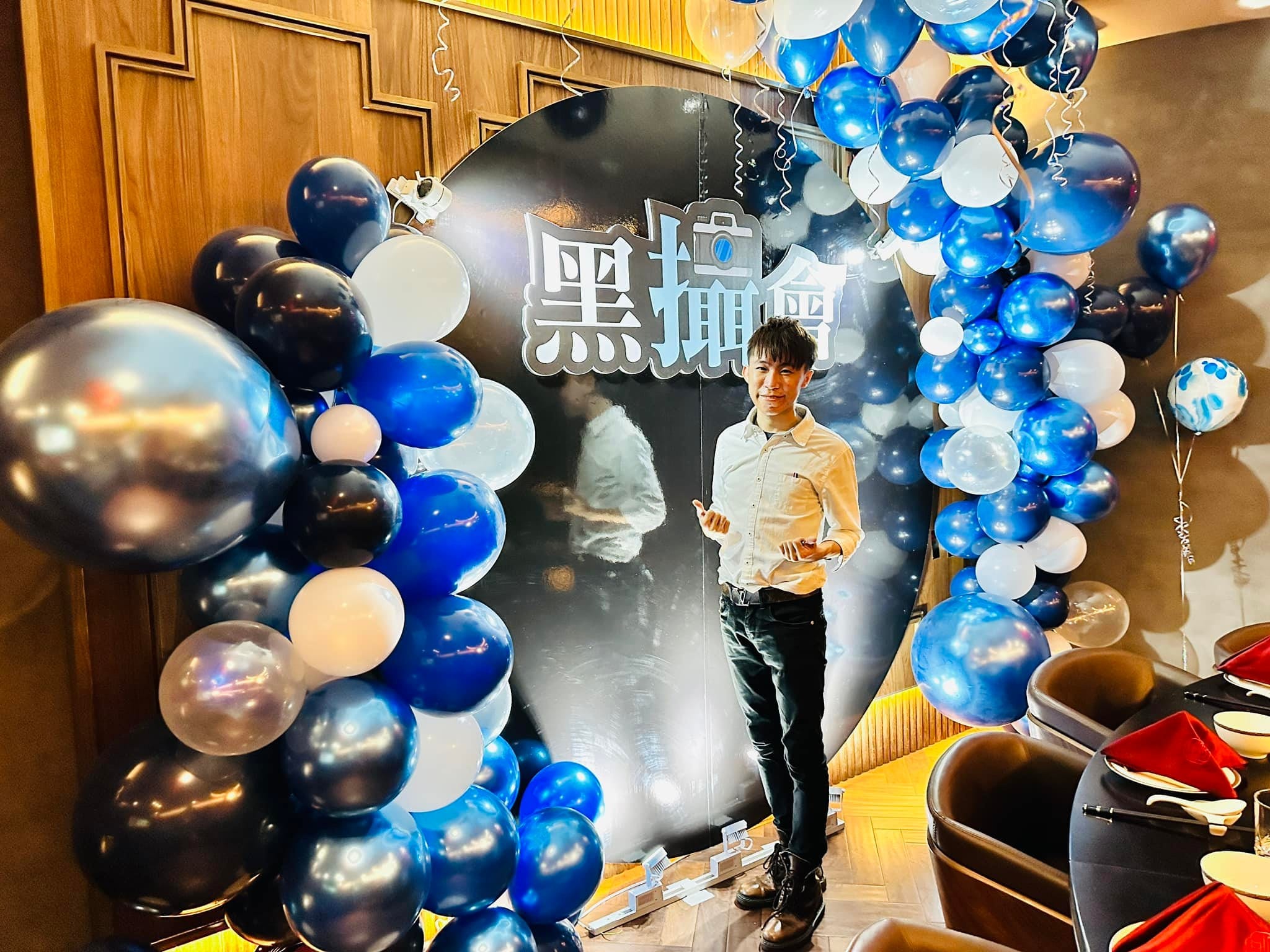 柯男站在黑色圓形背板前，左右是大小不一的藍色、銀色、白色氣球