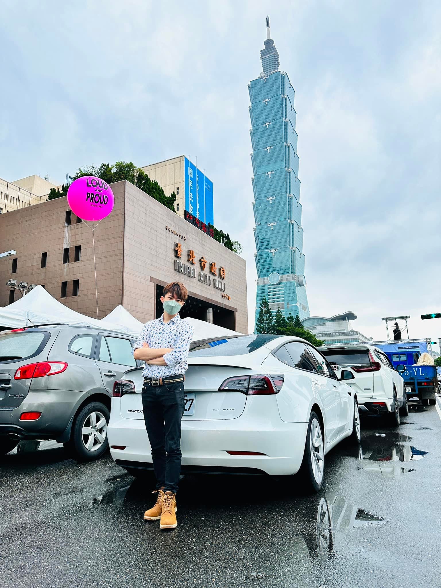 柯男站在台北市政府旁車邊，後方是台北 101 與桃紅色氣球