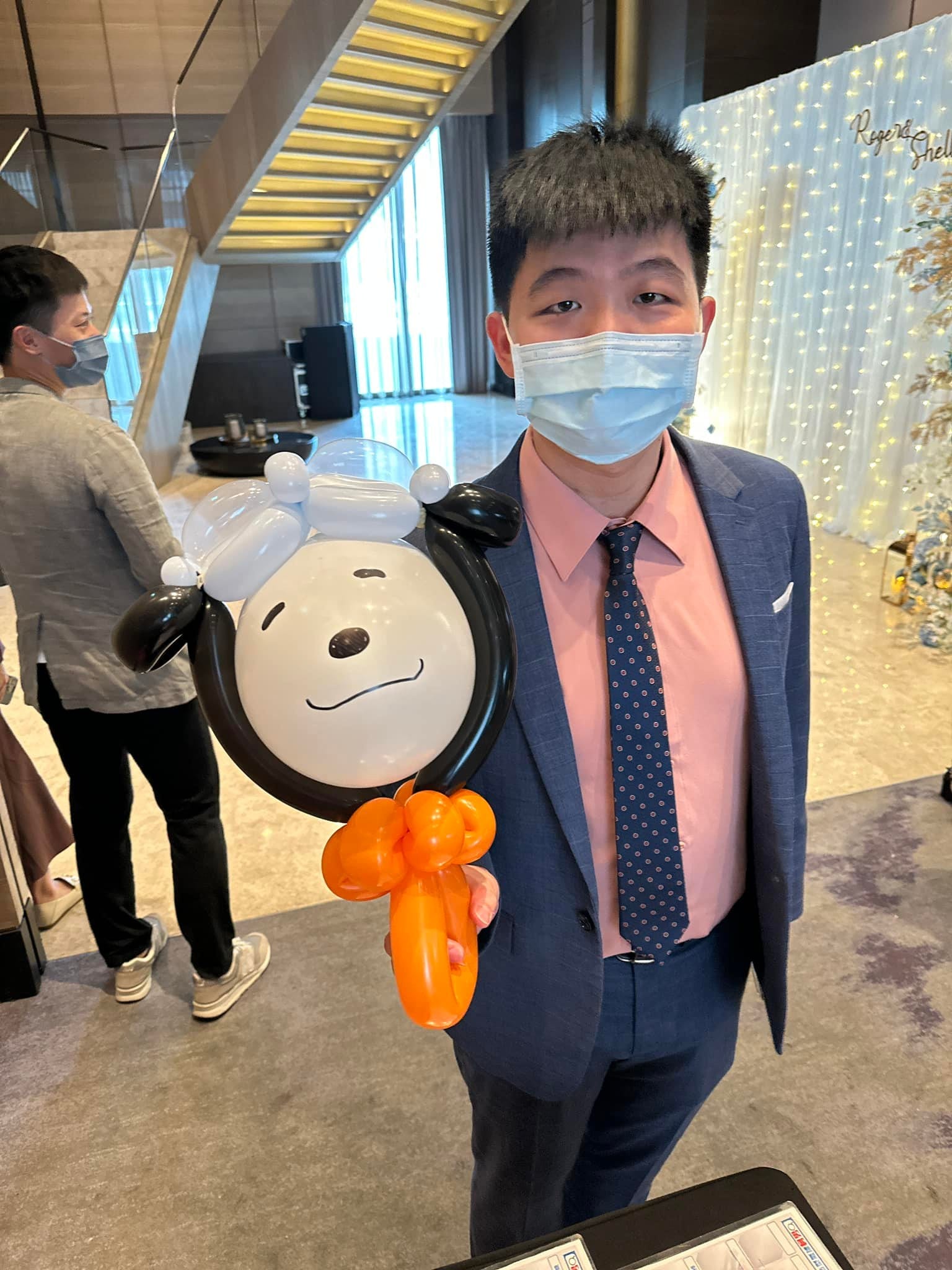 穿西裝的男賓客拿著黑耳白臉、橘色蝴蝶結的小狗造型氣球，背後是寫著新人名字的燈串背板