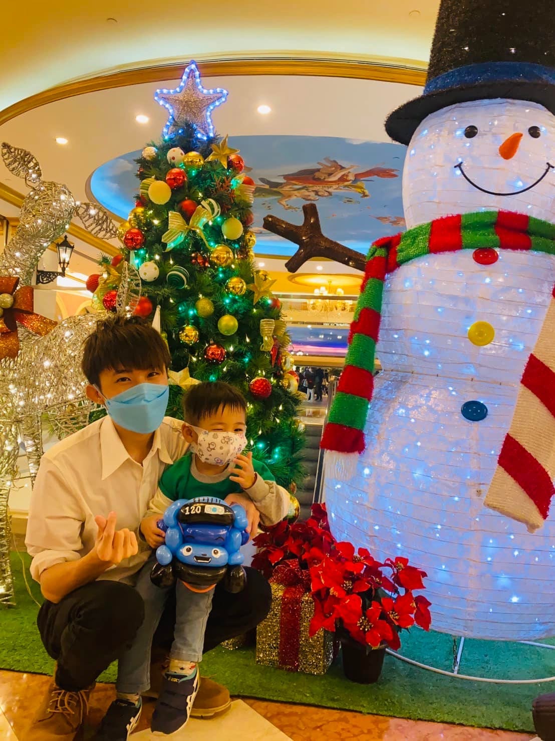 柯男蹲在大遠百聖誕樹與大雪人前，抱著拿 TAYO 小巴士氣球的小男孩