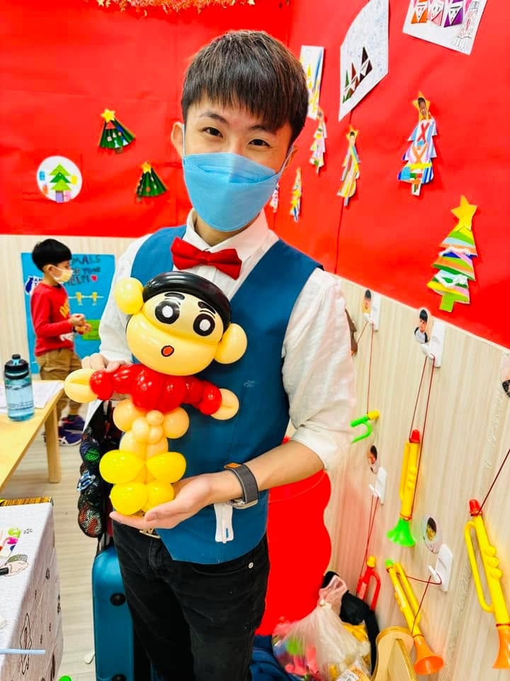 柯男手拿紅衣黃褲的蠟筆小新造型氣球，背景是紅色聖誕佈置牆