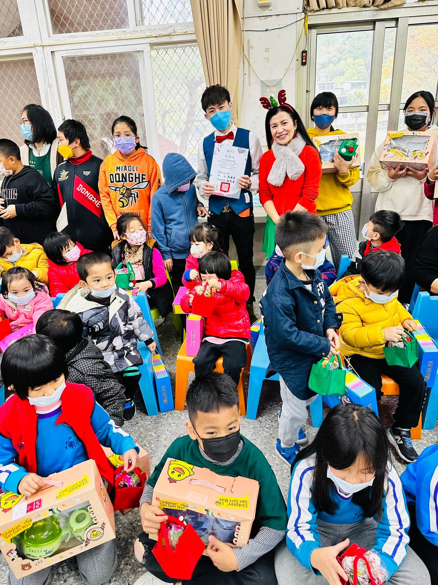 東河國小孩子們抱著聖誕禮物盒坐在彩色椅子上,柯男在後方拿著感謝狀