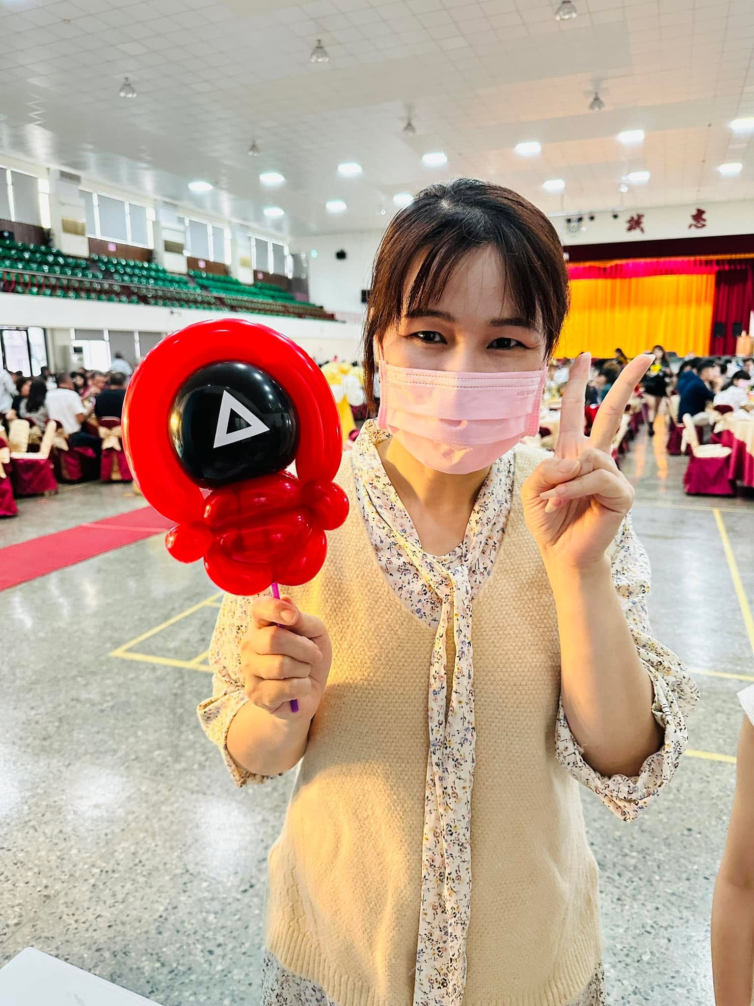 女賓客比 YA，手拿紅色兜帽、黑臉白三角形的魷魚遊戲面具氣球
