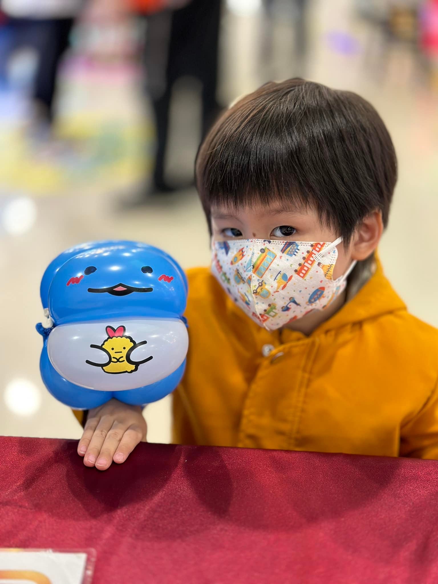 穿黃色外套的男孩拿著藍色圓胖、肚子有黃色圖案的造型氣球