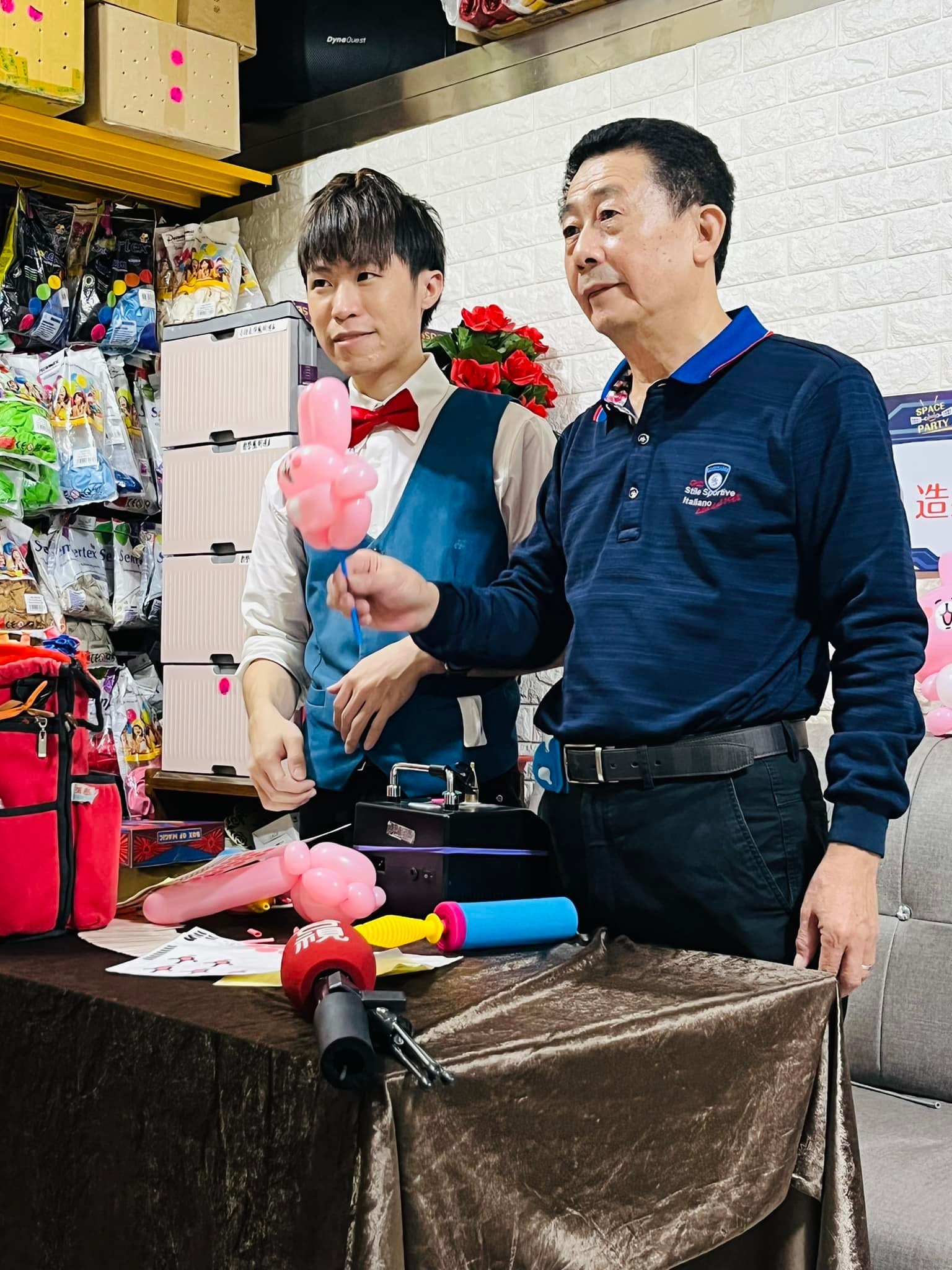 柯爹拿著粉紅氣球示範，柯男站在後方，桌上擺著採訪麥克風