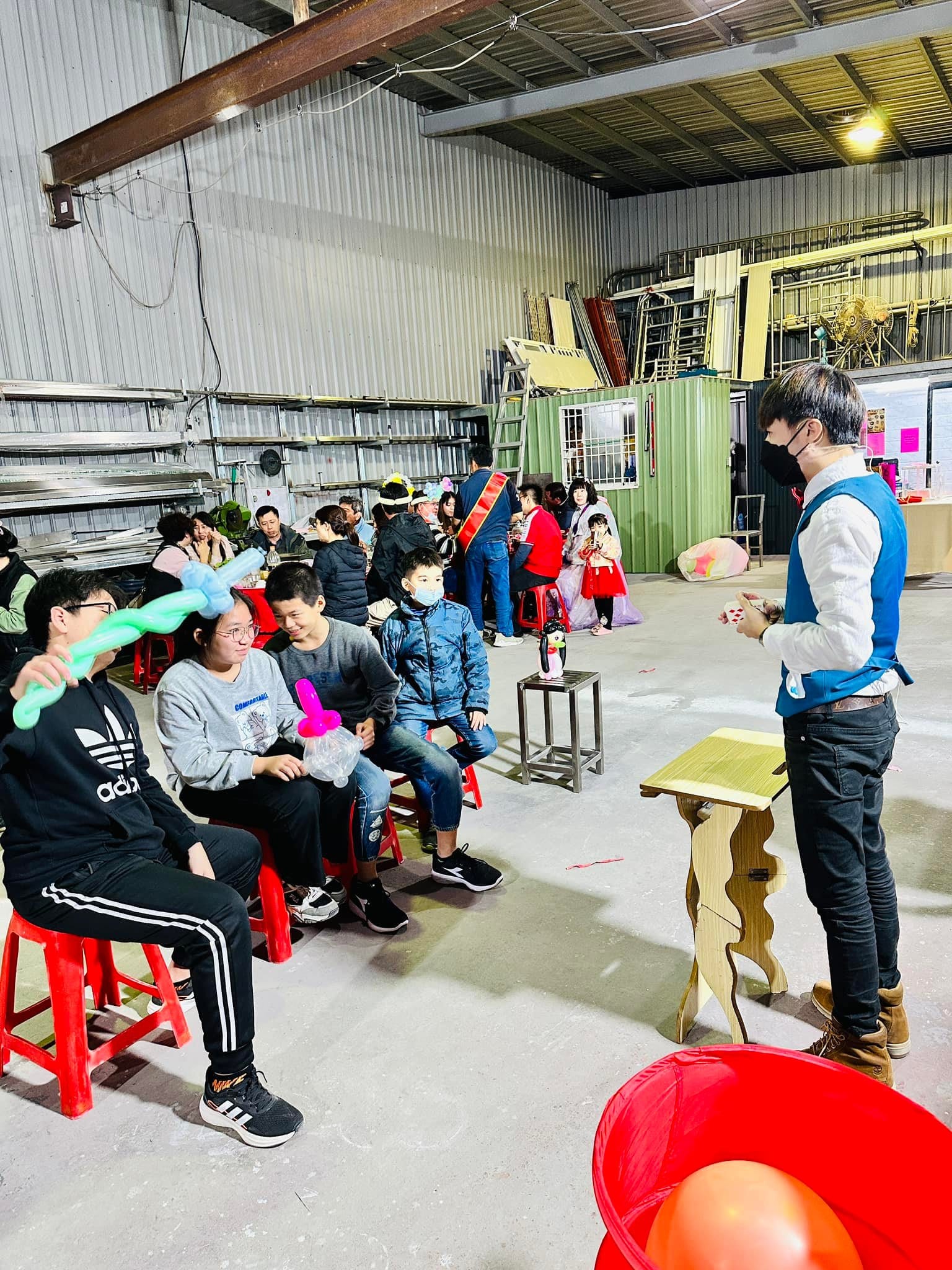 倉庫改成的尾牙會場，柯男站在道具桌旁表演魔術，賓客坐在紅椅上觀看