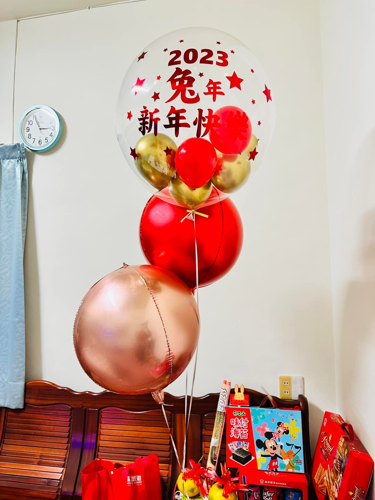 老家客廳的木椅旁，兔年新年快樂波波球下方串著紅色與玫瑰金圓形鋁箔氣球，椅子上放著年節禮盒