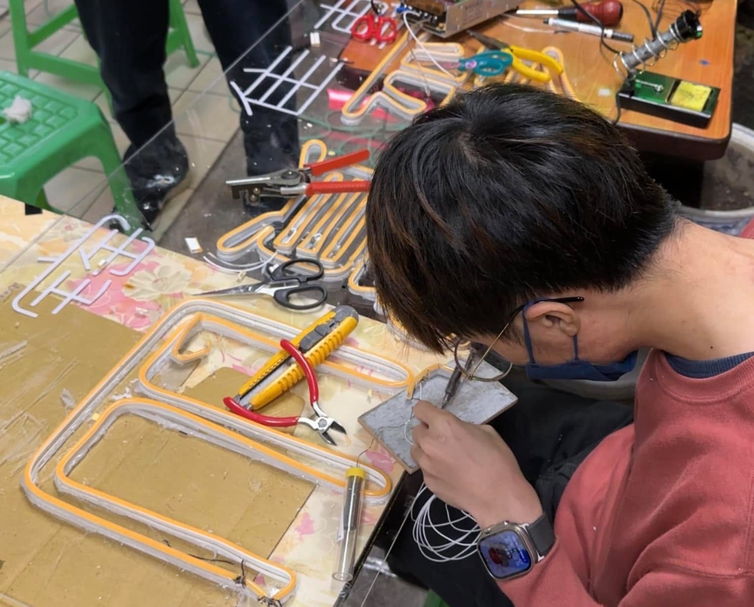 柯男低頭焊接 LED 燈條線路，桌上放著鉗子與壓克力板