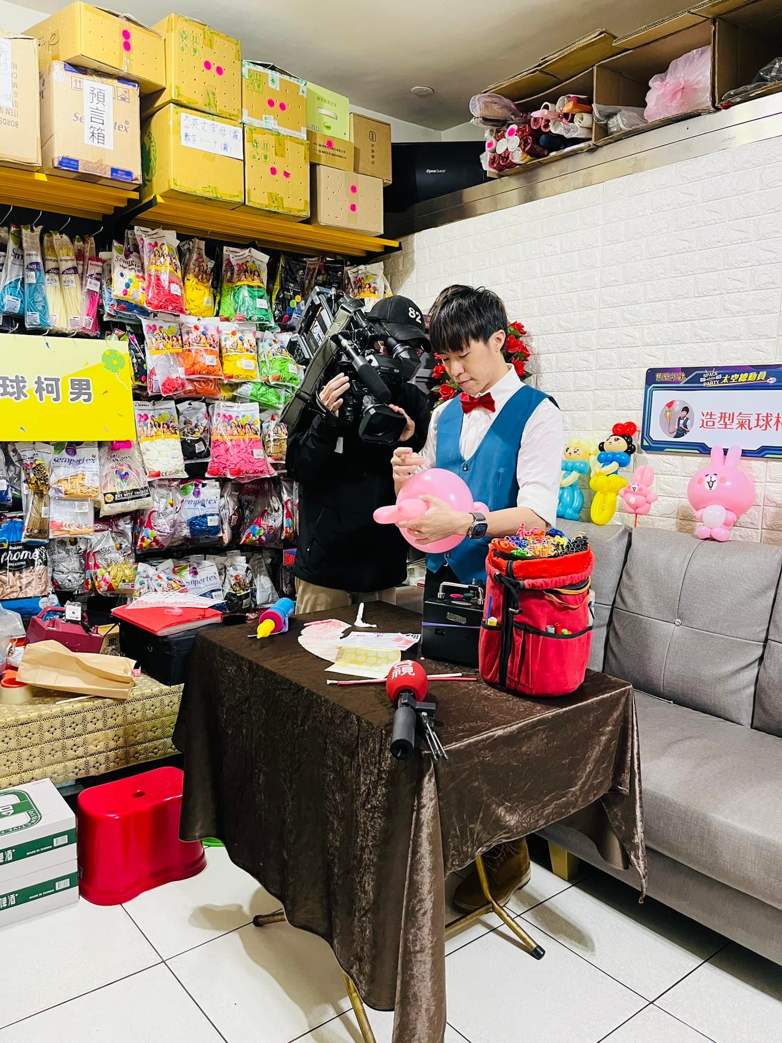 台視攝影師扛著攝影機，拍攝柯男坐在桌前折粉紅色氣球