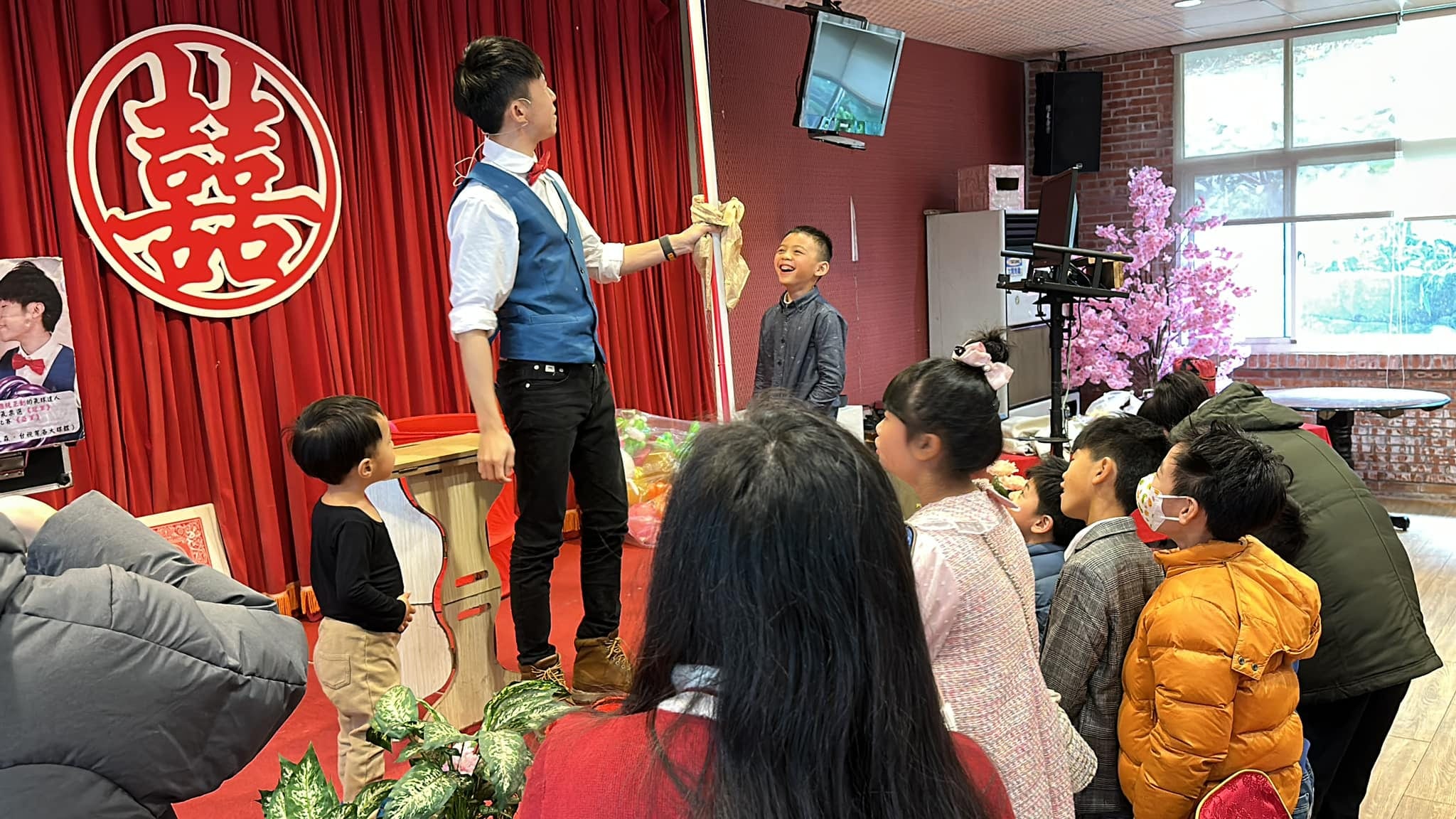 紅色囍字背景前，柯男拿著長桿道具，小男孩站在舞台上當助手