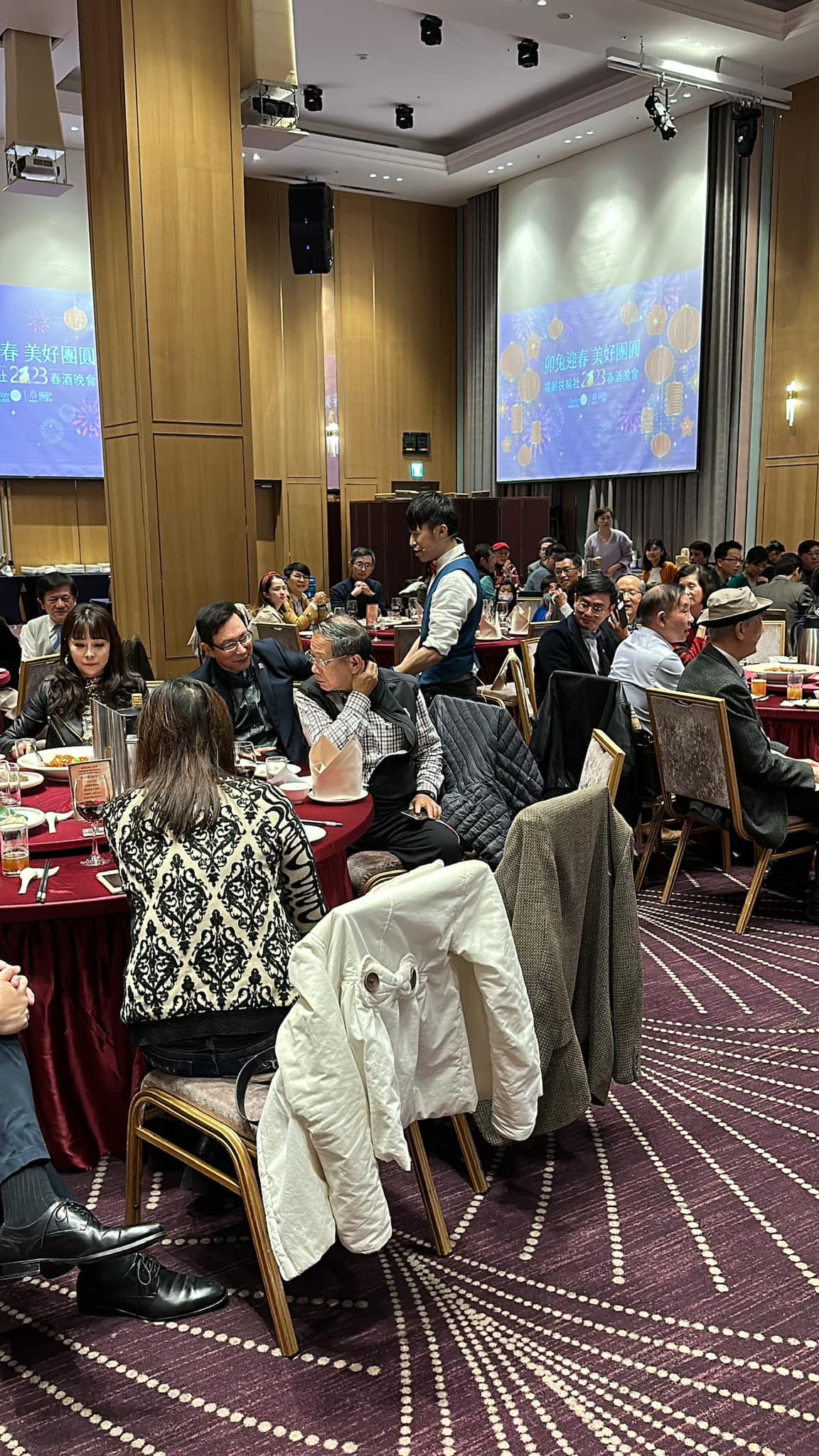 春酒晚會圓桌之間，賓客用餐，表演者在後方桌邊服務