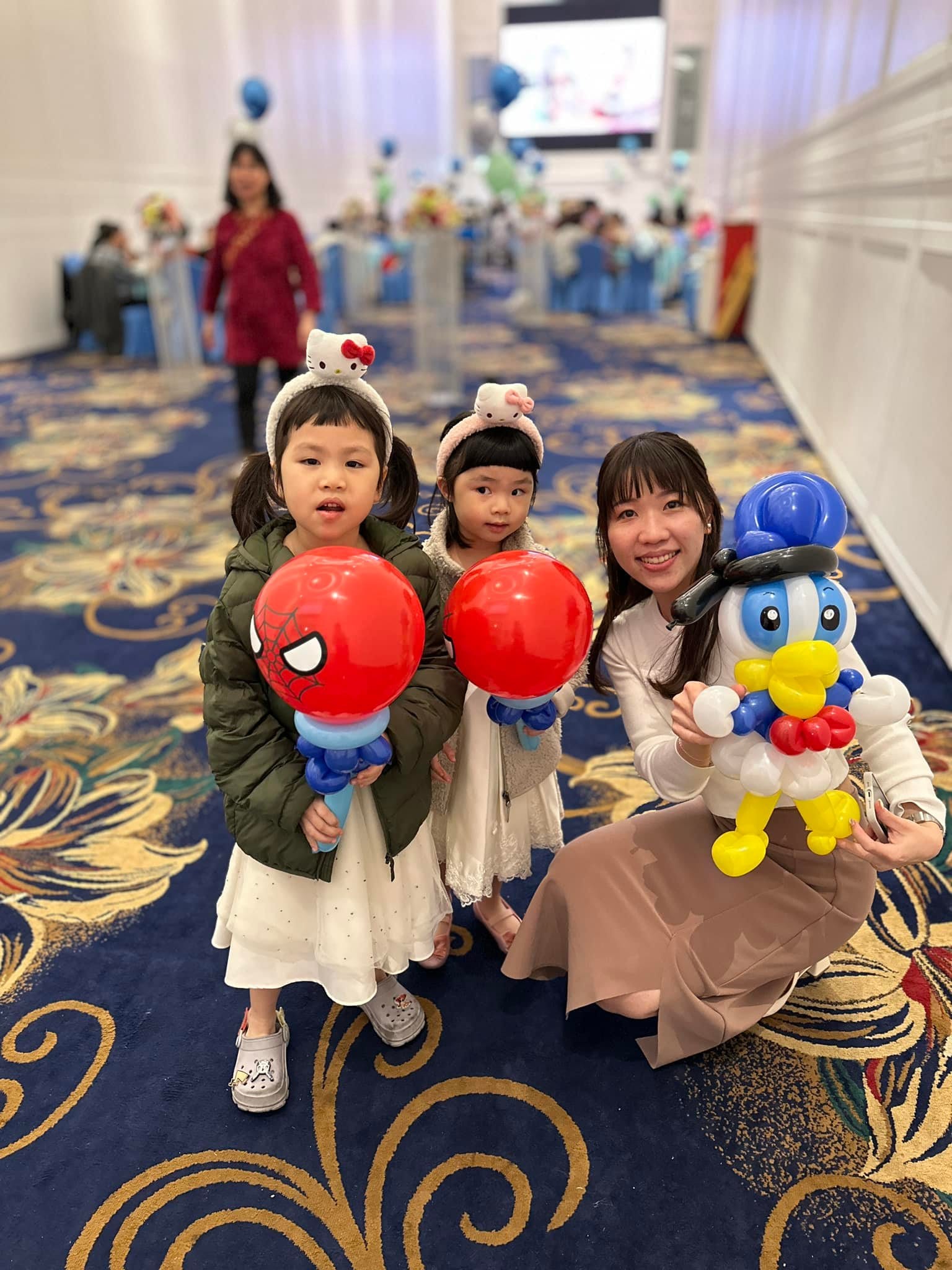 兩個小女孩拿著蜘蛛人頭氣球棒，媽媽抱著唐老鴨氣球蹲在一旁
