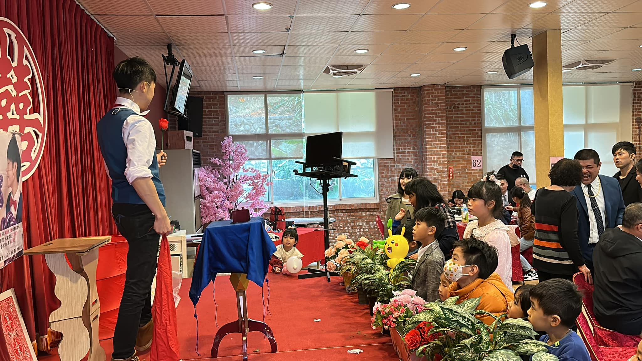 婚宴會場裡，賓客與孩子們坐著看柯男在藍色道具桌旁表演