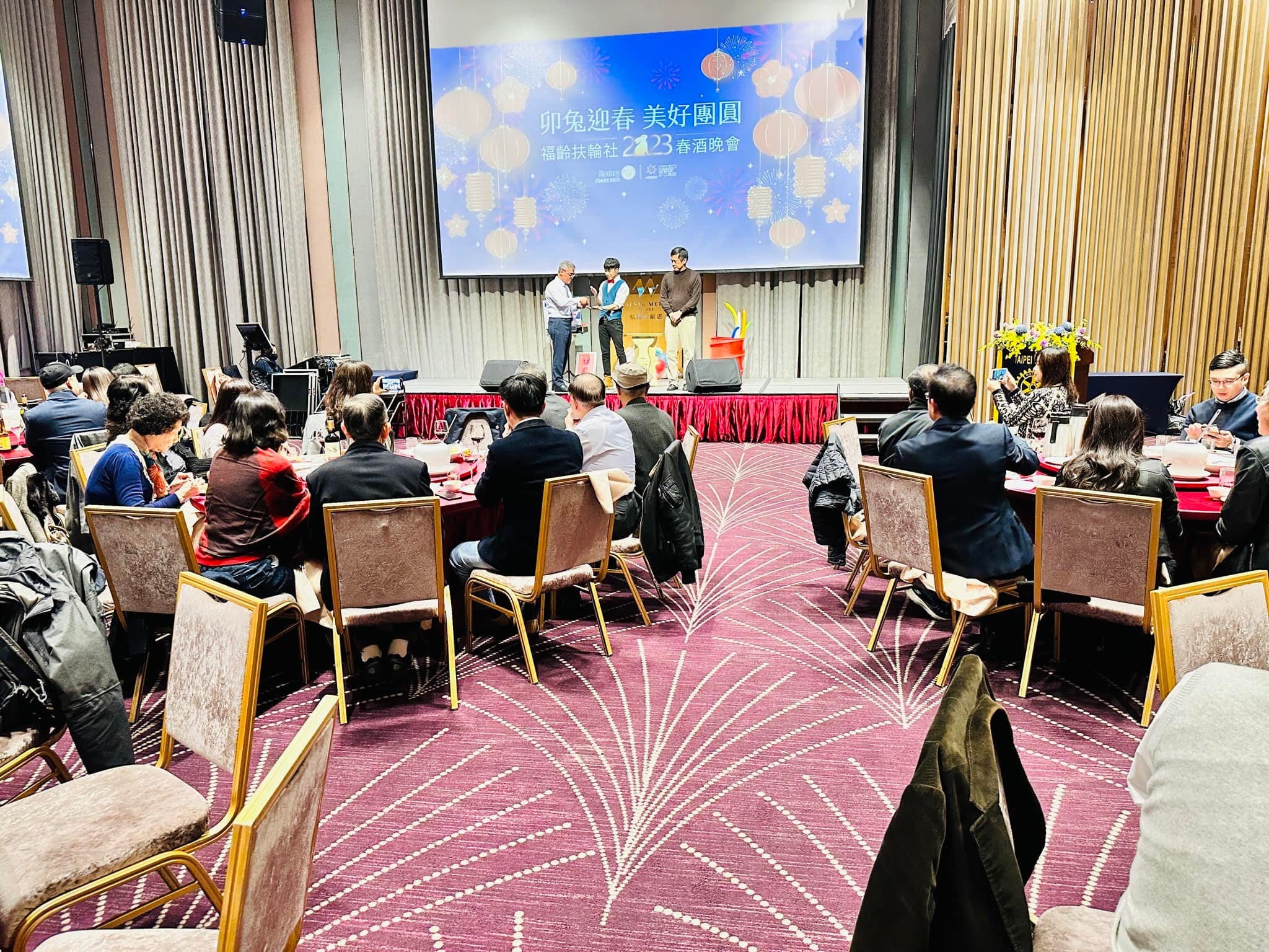 扶輪社春酒晚會宴會廳，賓客圍桌而坐，前方舞台與投影幕