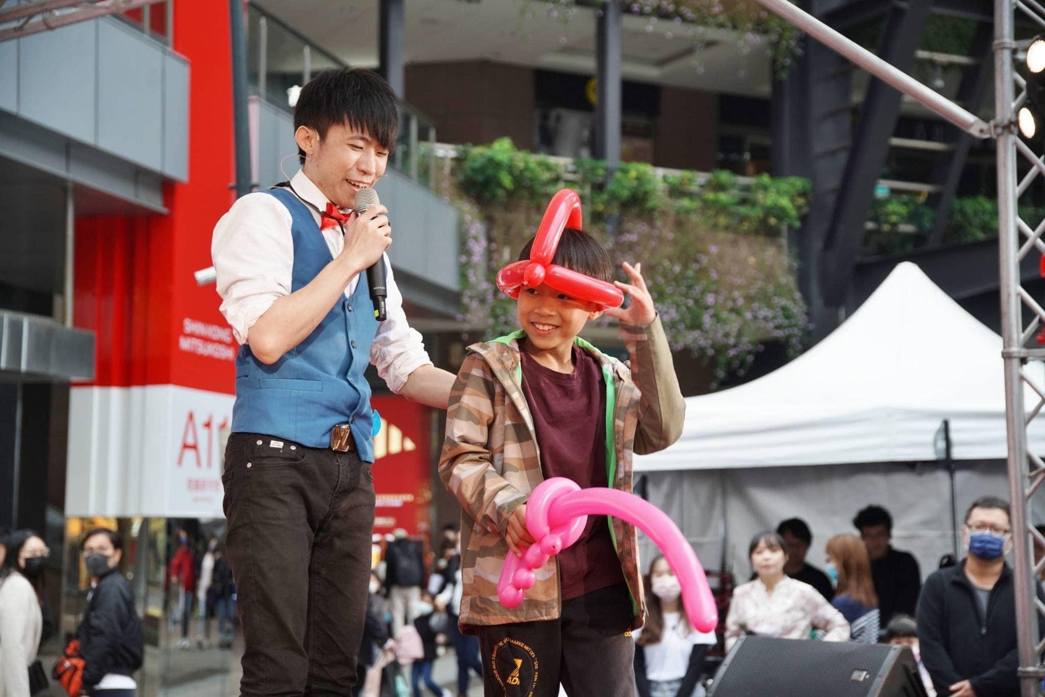 上台的男孩戴著紅色氣球帽、手拿粉紅氣球，和柯男一起表演