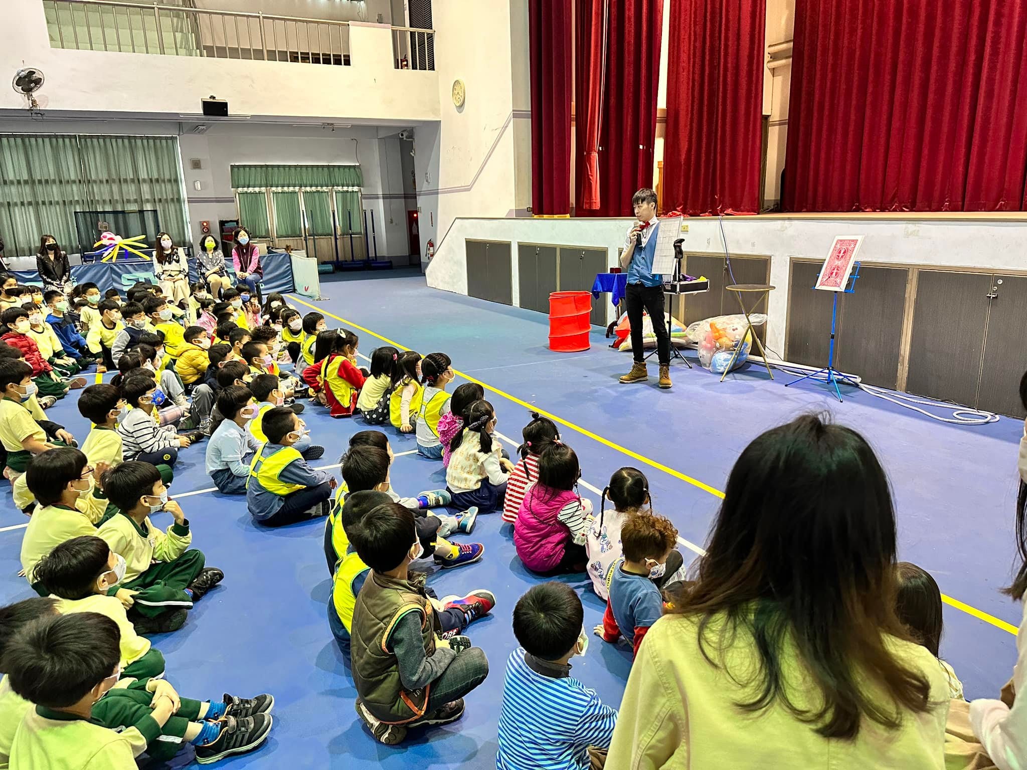 柯男在活動中心舞台前拿麥克風表演，孩子們席地而坐專心看