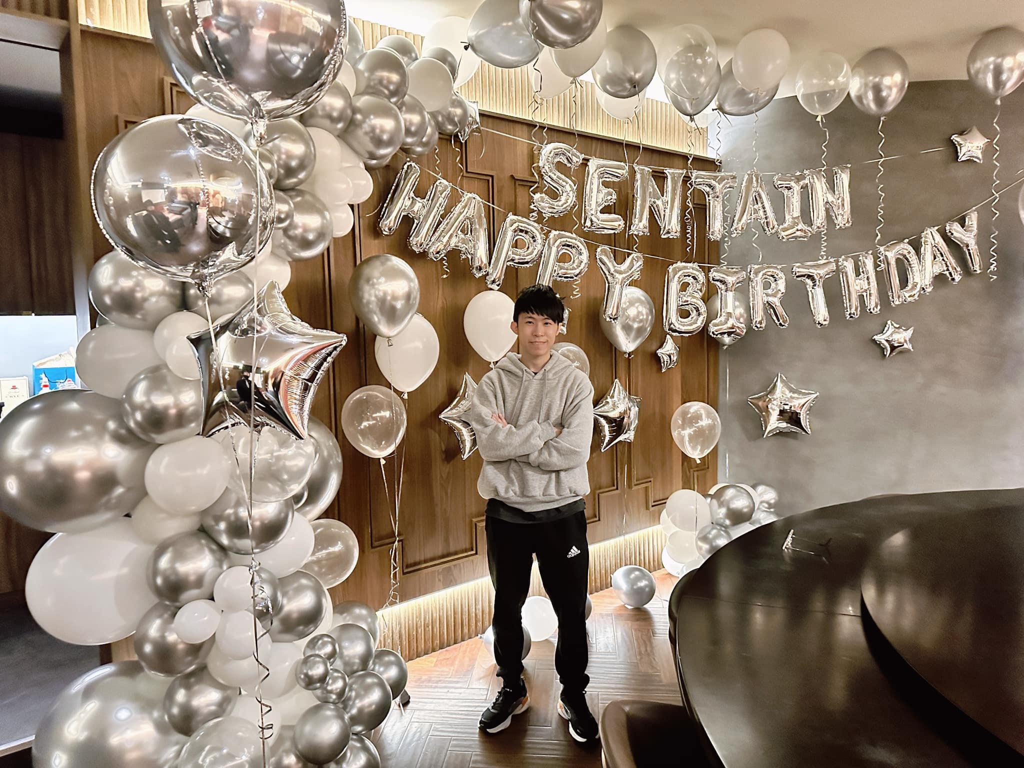 柯男雙手抱胸站在銀白色氣球串與 HAPPY BIRTHDAY 字母氣球前