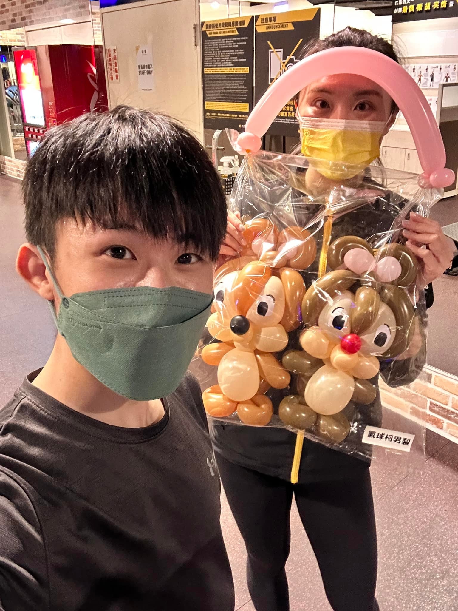 健身房裡，戴口罩的教練舉著奇奇蒂蒂氣球提袋與柯男自拍
