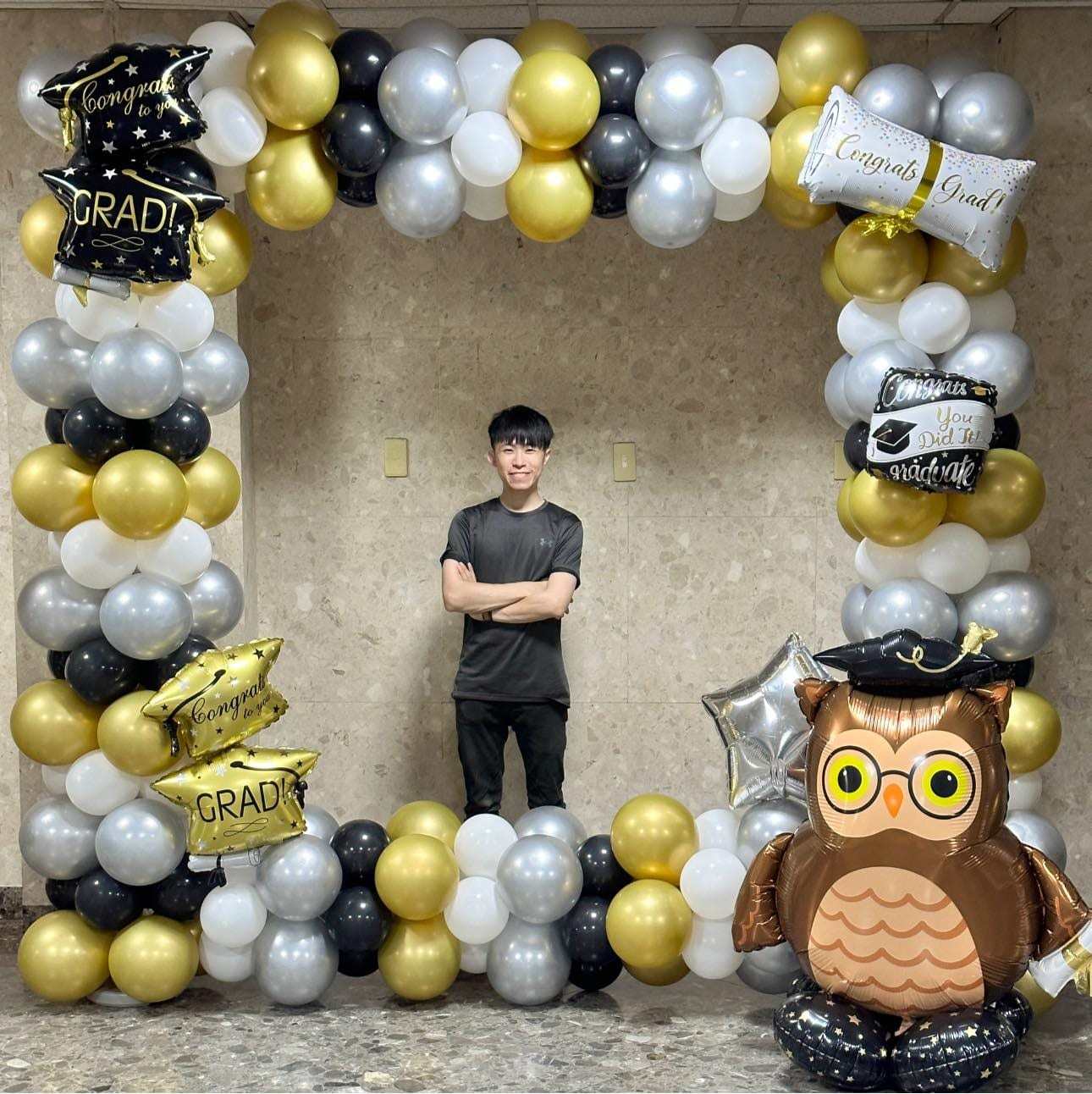 金銀黑白氣球圍成的方形畢業拱框，四周掛博士帽 GRAD 與畢業證書鋁箔氣球，右下角有戴博士帽的貓頭鷹氣球，我站在框中央