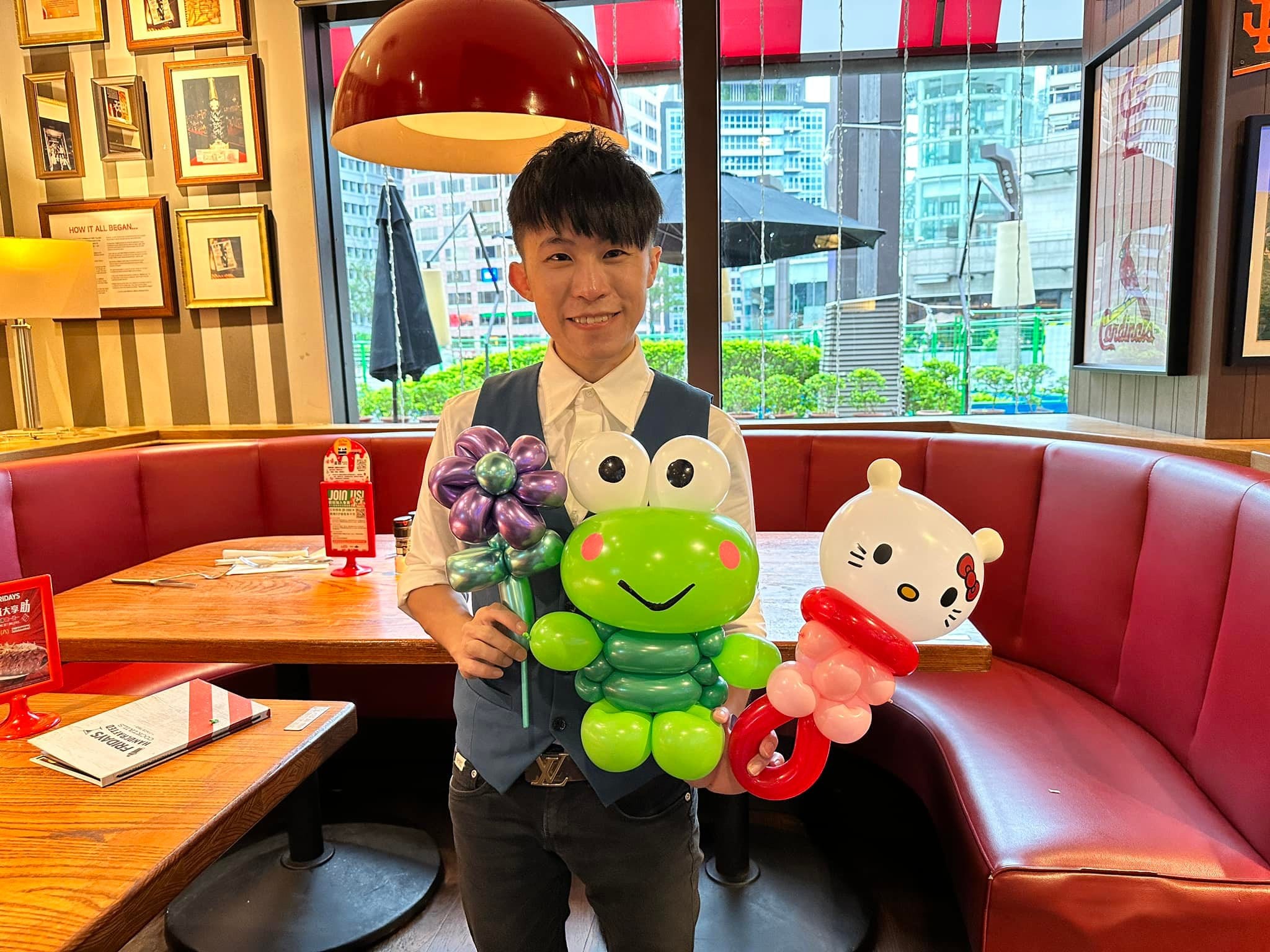 柯男在餐廳卡座前拿著紫色花朵、綠色青蛙和 Hello Kitty 造型氣球