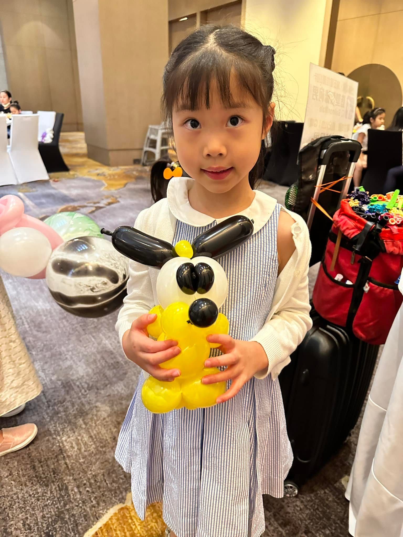 小女孩抱著黑耳朵的黃色小狗造型氣球，站在宴會廳裡