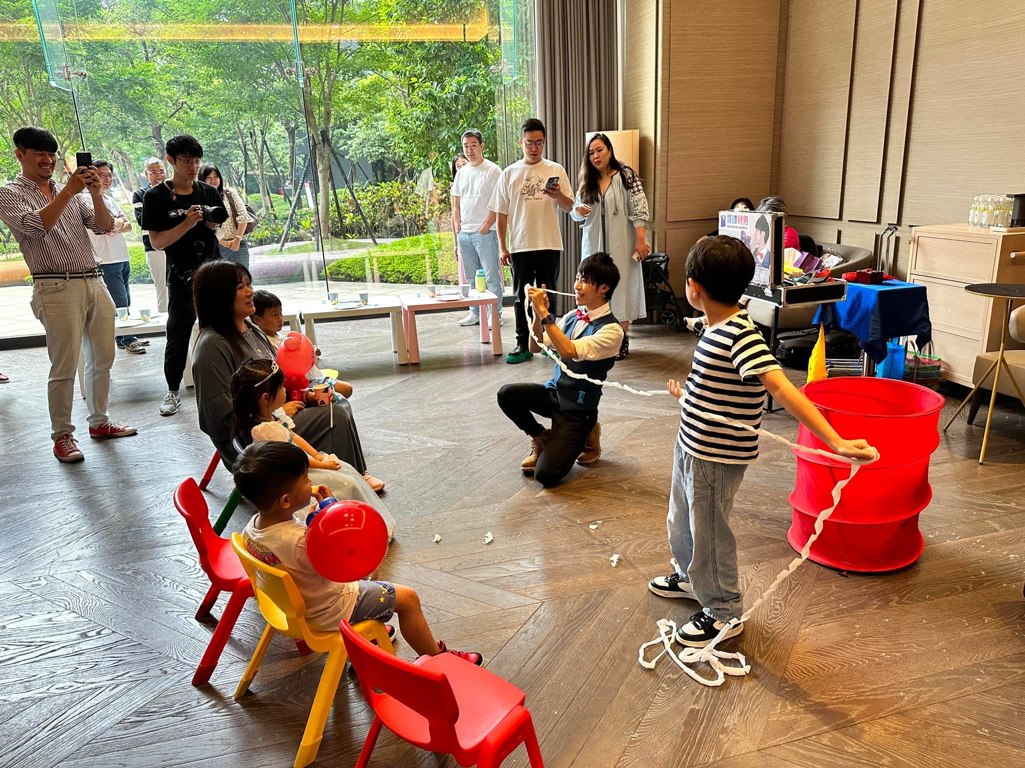 交誼廳裡柯男蹲著表演繩子魔術，男孩拉著繩子，孩子坐在紅色小椅子上