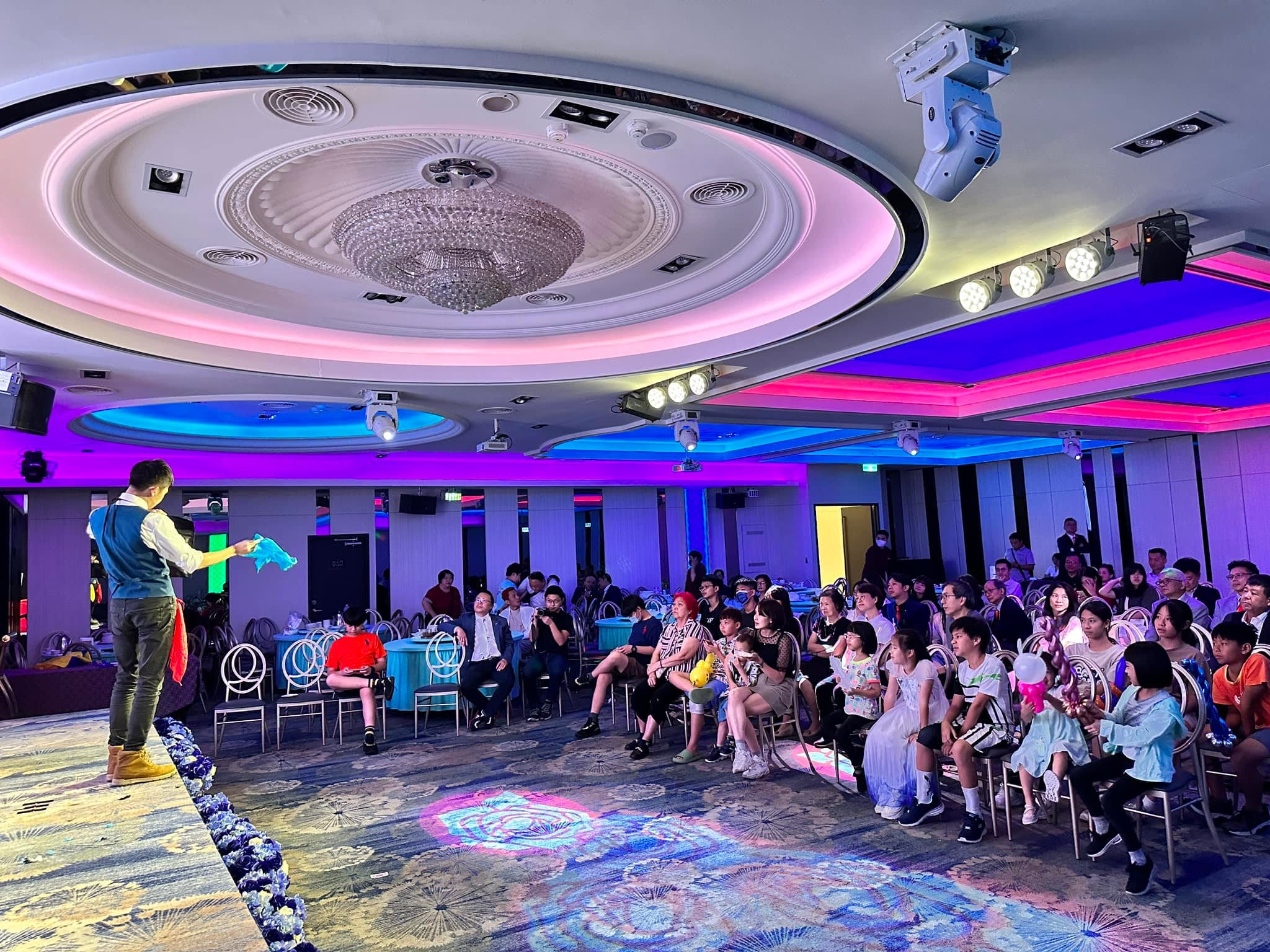 扶輪社例會在宴會廳舉行，柯男站在伸展台邊表演，社友與孩子坐滿觀眾席