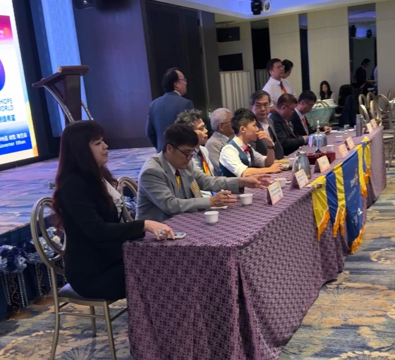 柯男穿表演背心坐在例會主桌，與西裝社友並排，桌上放著名牌與茶杯